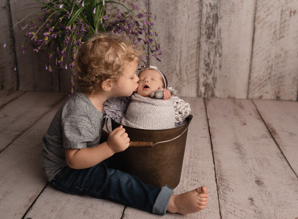 Toddler big brother nuzzling his newborn sibling tucked into a bucket during a Leander studio session