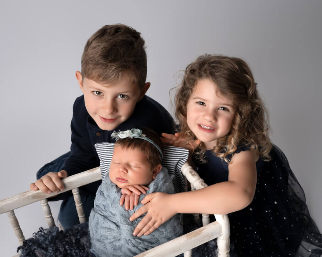 Big brother and sister holding their newborn sibling on a small bench in an Austin studio
