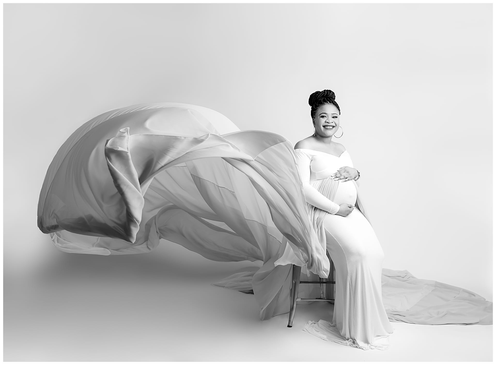 black and white photo of a woman sitting holding her pregnant belly with a dress flying around her.