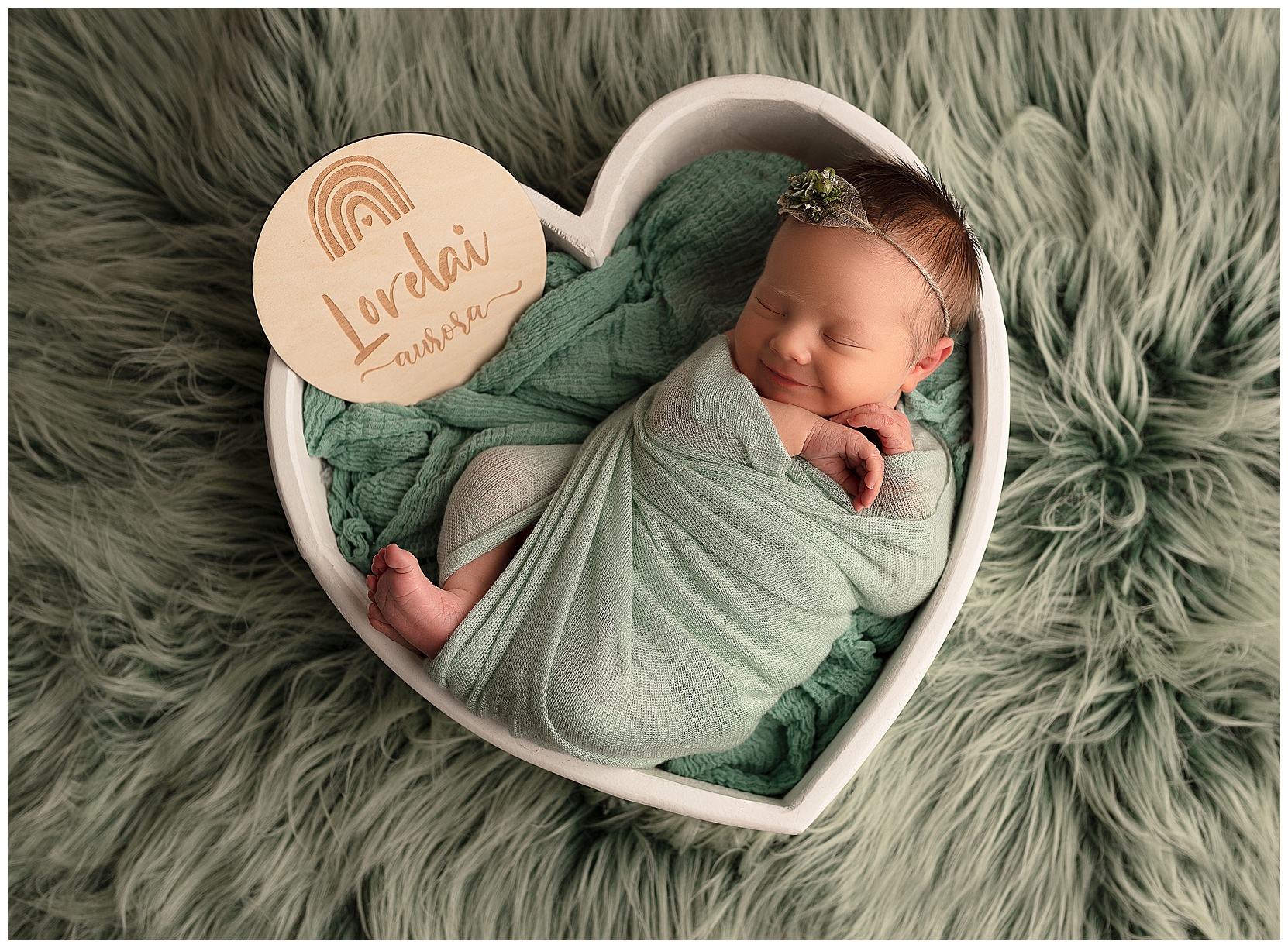 smiling newborn baby in a sea green wrap sleeping in a heart bowl
