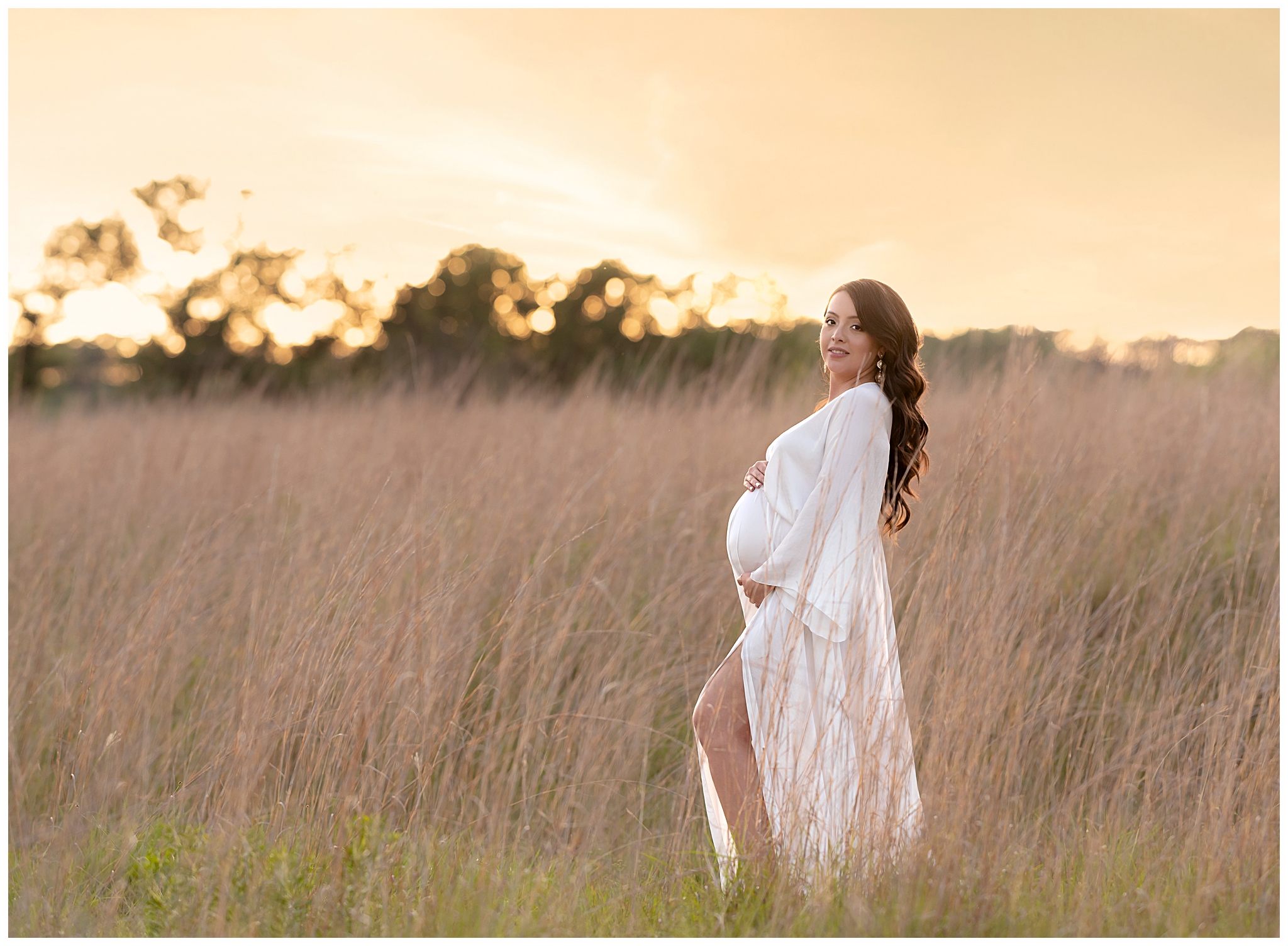 Pregnant woman in a white maternity gown stands in a field of long grass in front of a sunset