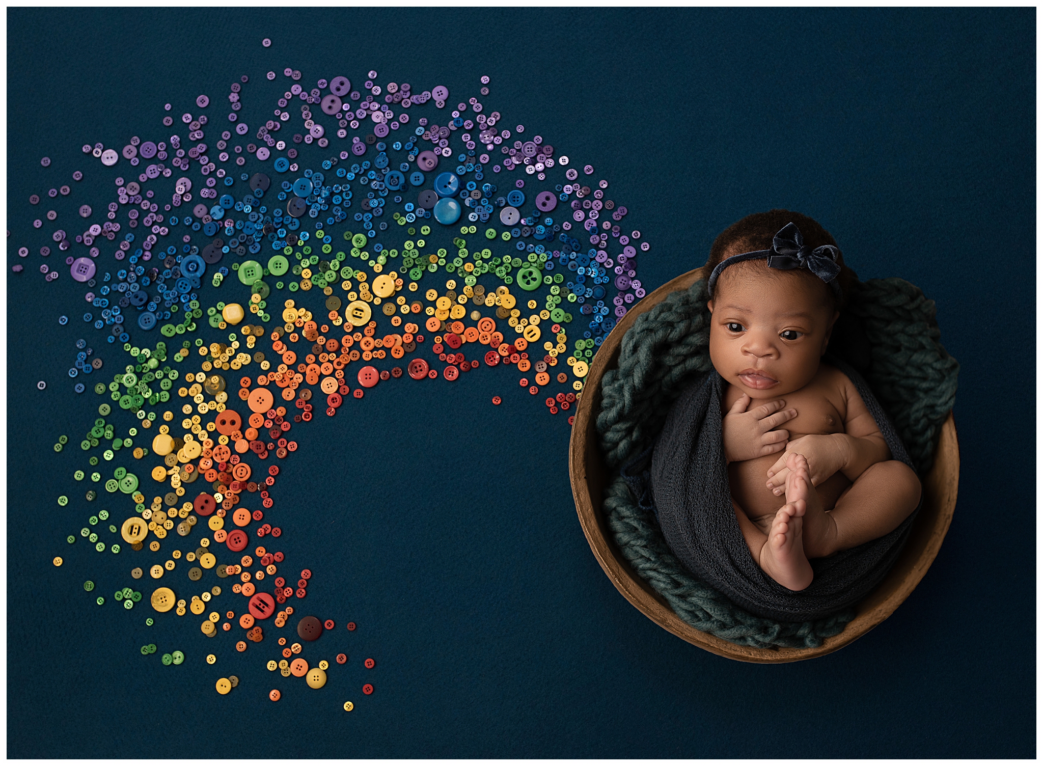 Newborn baby with big brown eyes and brown skin wearing a bow, in a basket, on a rainbow background