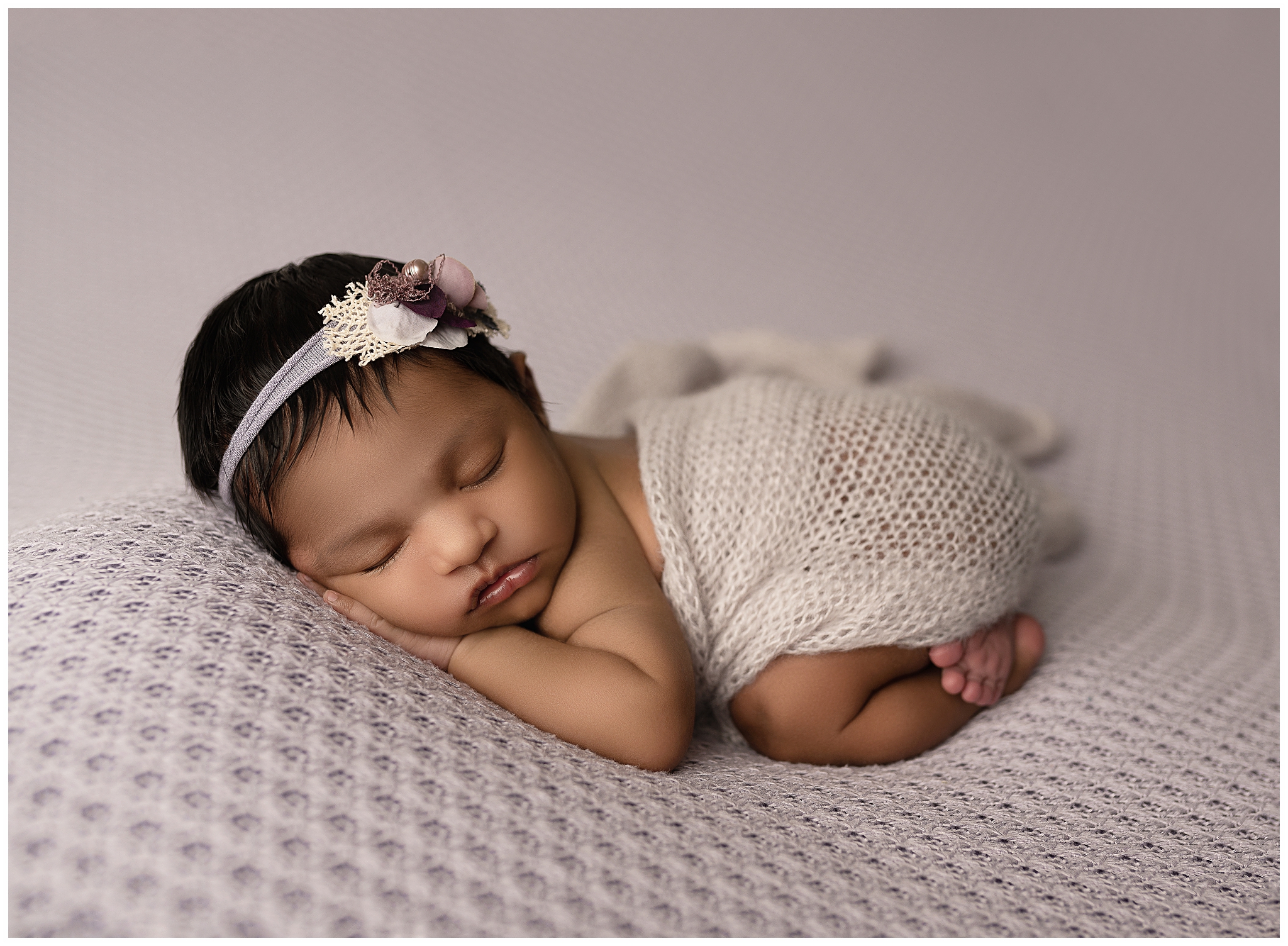 Sleeping baby on her belly laying on a purple blanket wrapped in a purple knit fabric and wearing a purple headband.