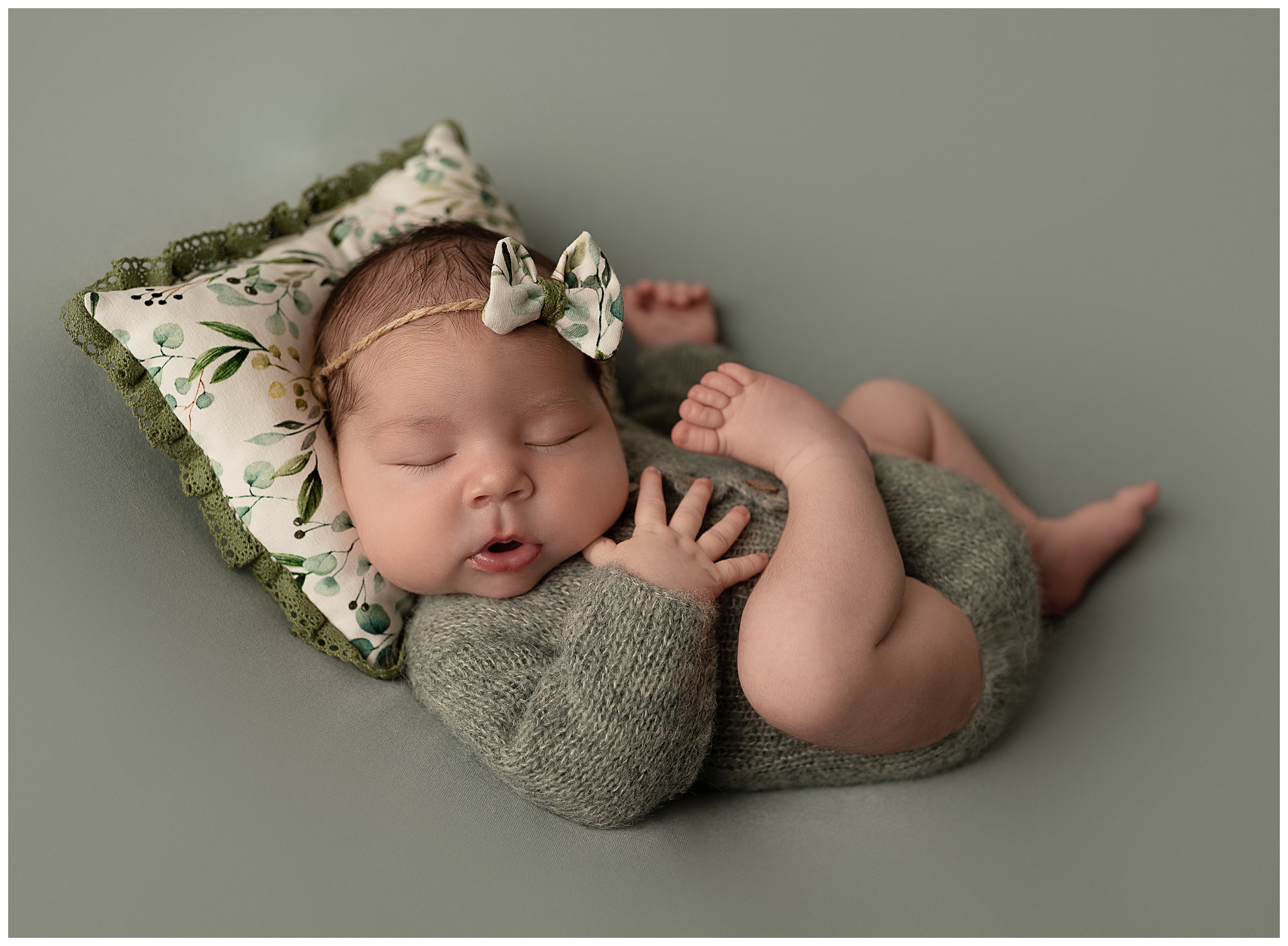 Baby wearing green knit romper sleeping on her back laying on green floral pillow with green floral bow on her head