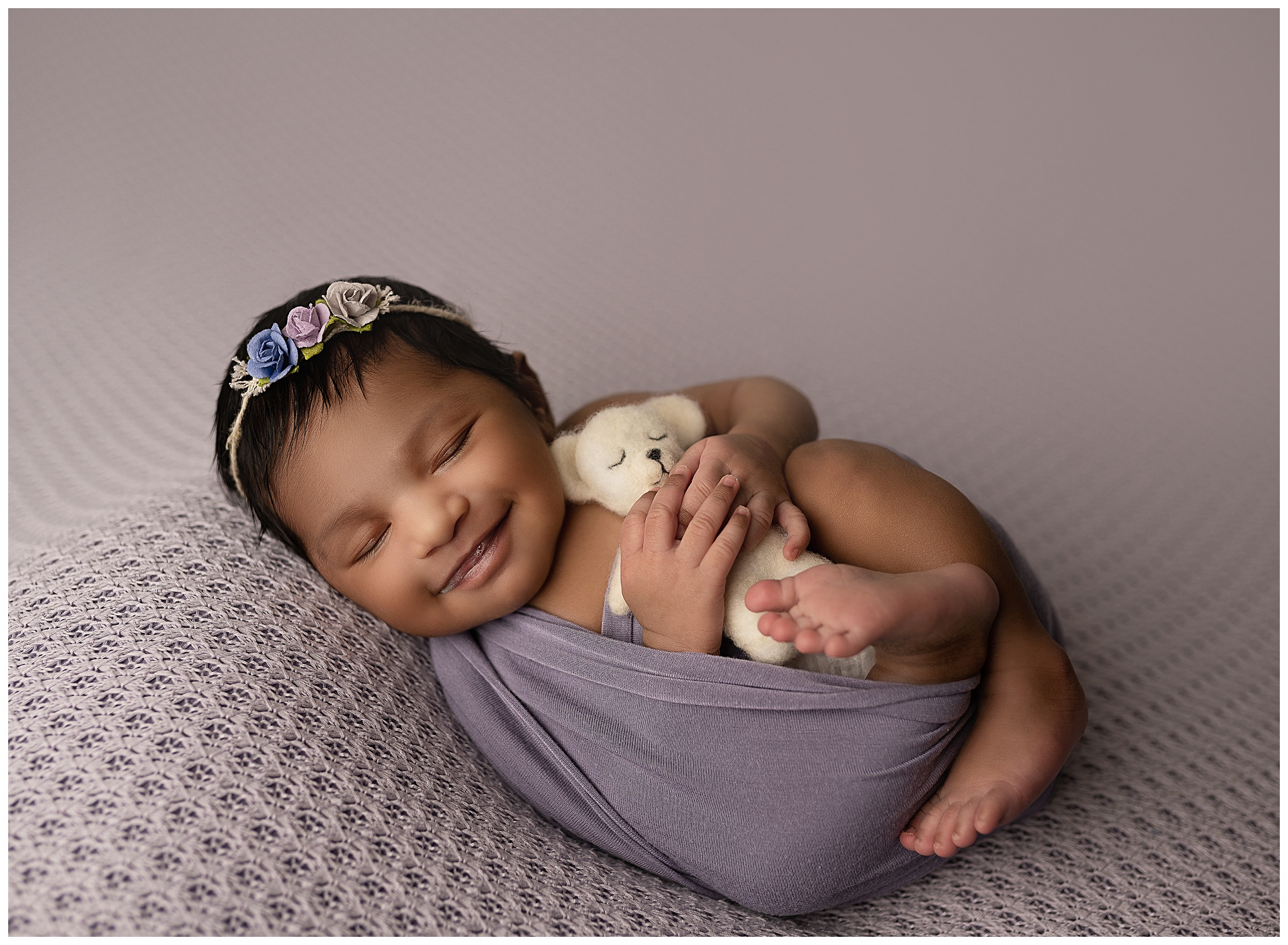 Smiling newborn baby wrapped in purple holding a white teddy bear.
