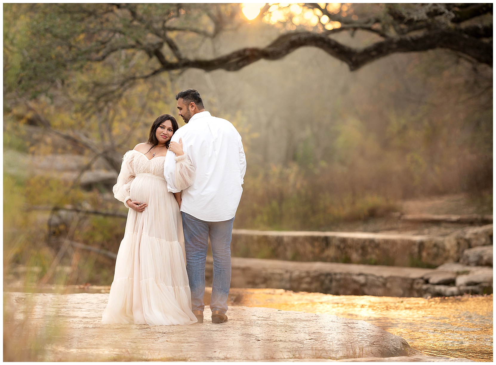 Austin TX maternity photo with Bull Creek in the background. Features a woman in a long, white dress facing the camera. She is holding the arm of her husband, who is wearing a white button-down shirt and jeans. He is facing away from the camera, with his head tilted sidewards toward her. 