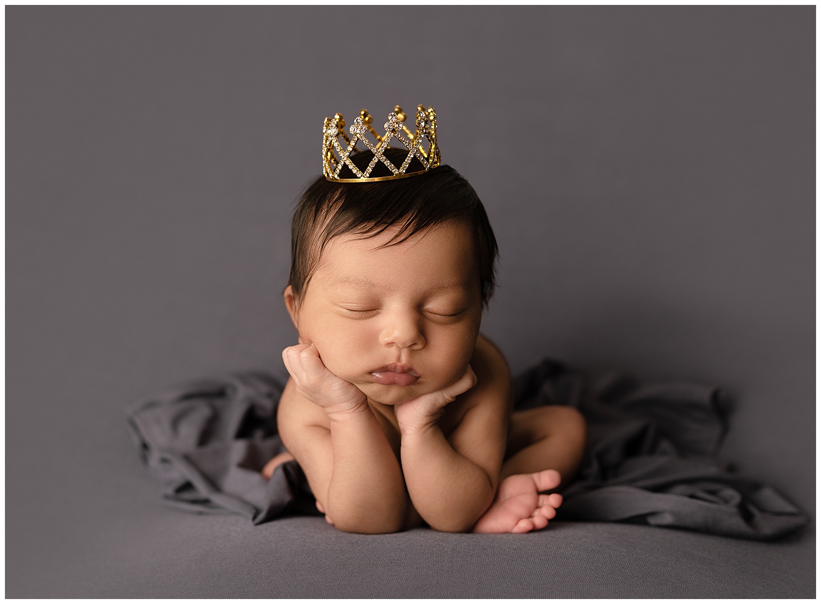 Newborn photo featuring a baby girl on a gray background. She is posed in the froggy pose with a small crown on her head.