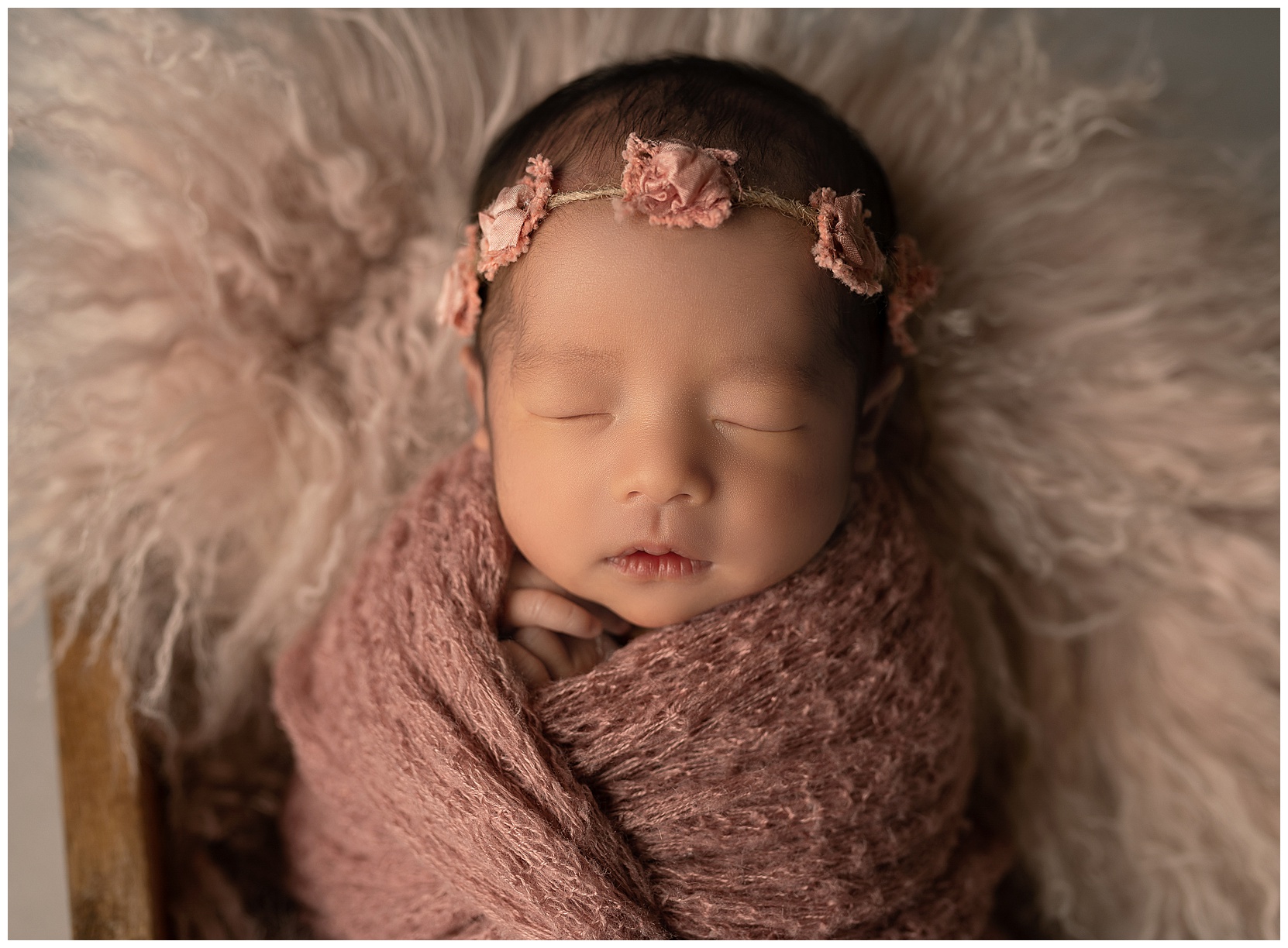 Close-up of a newborn wearing a pink flower crown, wrapped in a soft pink knit shawl on a sheepskin background