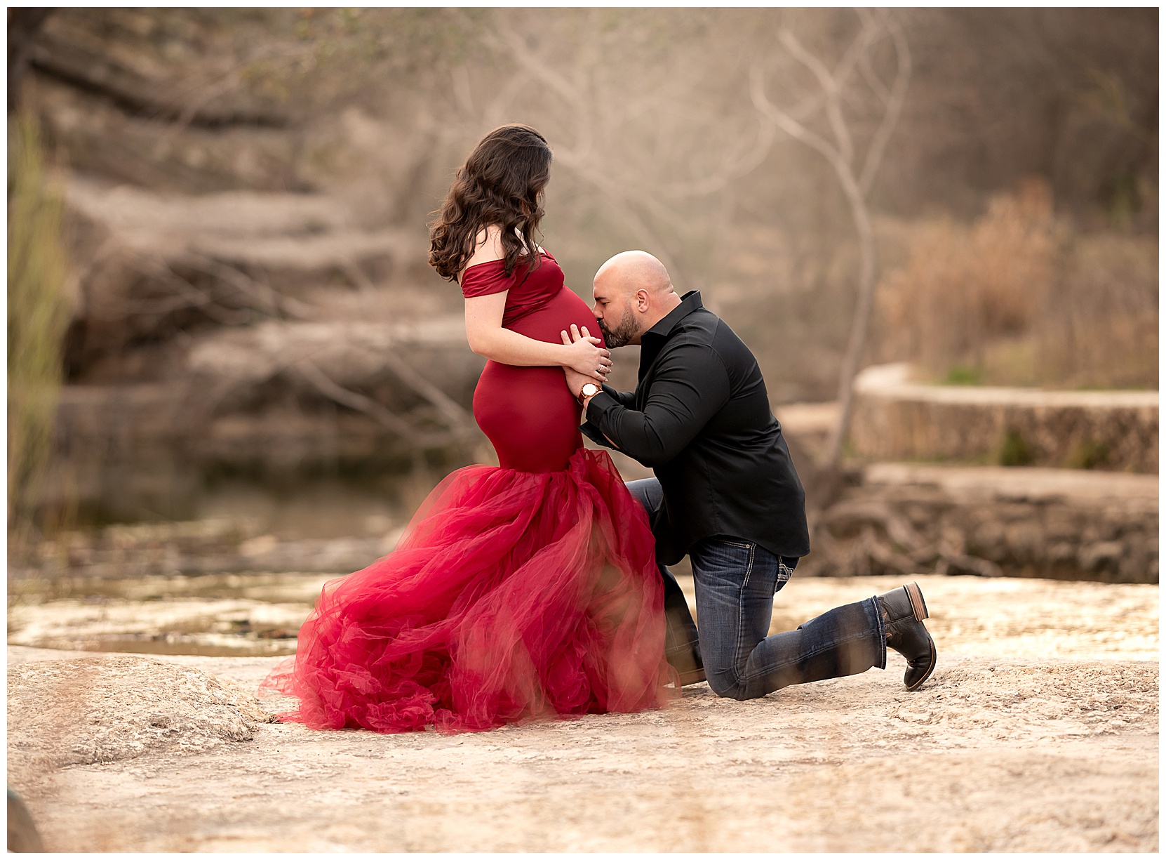 outdoor maternity photo picture featuring a woman in a red maternity gown, her partner is kneeling down in front of her and kissing her belly