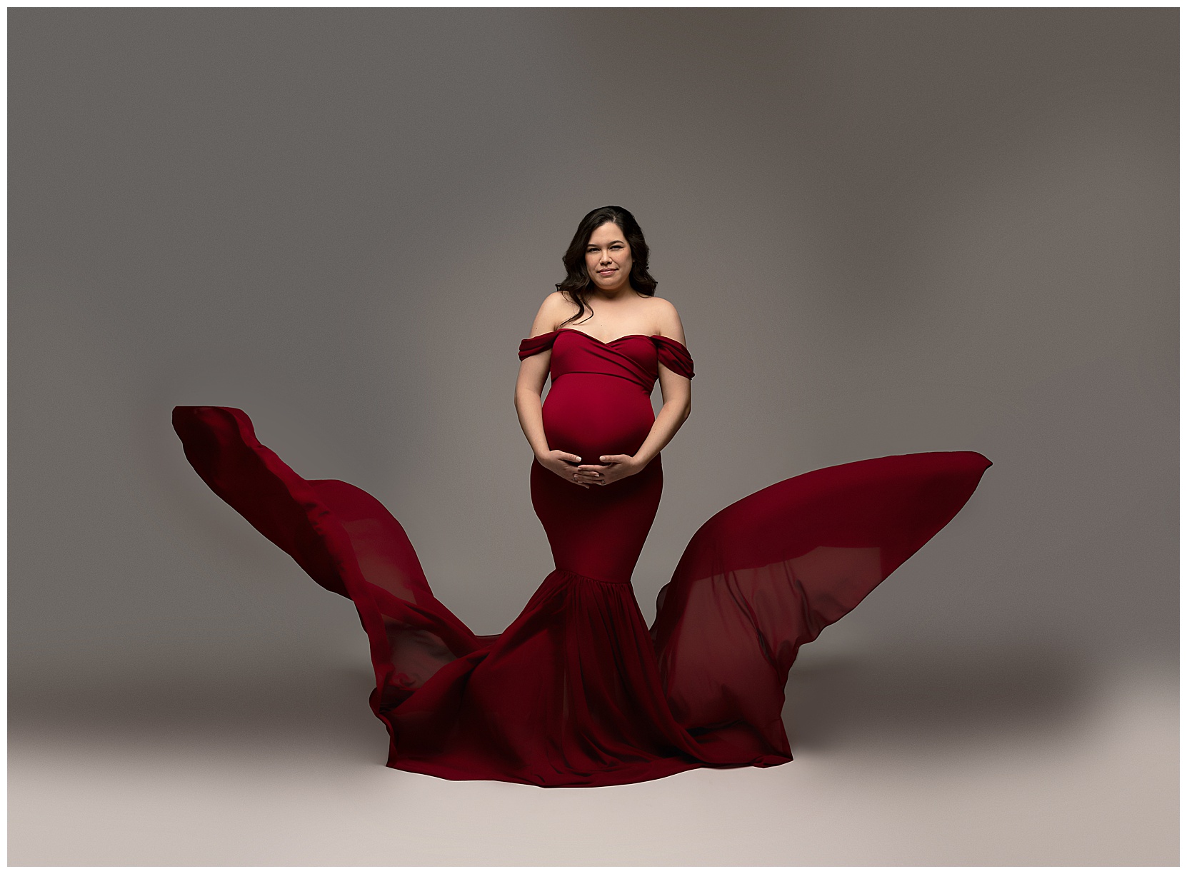 Unique maternity photo featuring a woman in a red dress cradling her belly. The train of her gown is floating on either side of her.