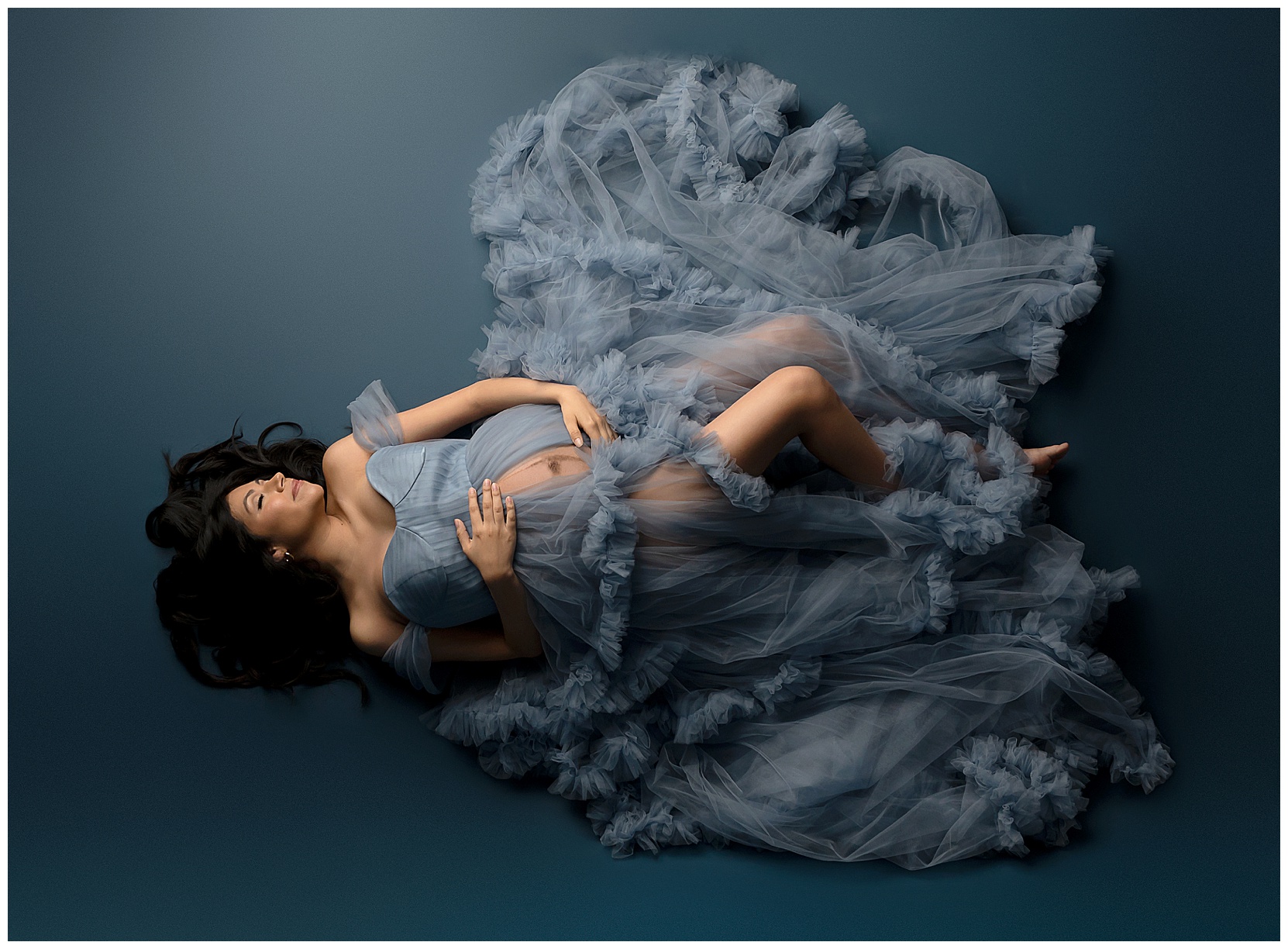Photo of a pregnant woman in the maternity studio. Woman is lying on her back on a blue backdrop, wearing a ruffled, dusty blue maternity gown.