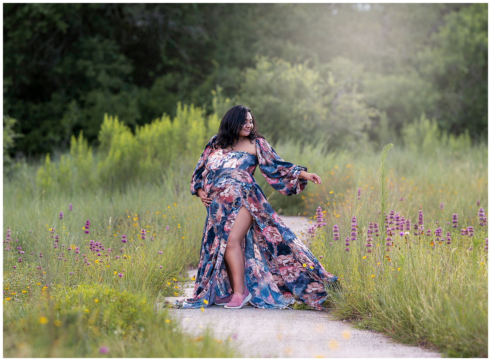 A color Texas wildflower maternity photo featuring a woman in a blue and pink floral dress and pink sneakers standing in a walkway surrounded by wildflowers.