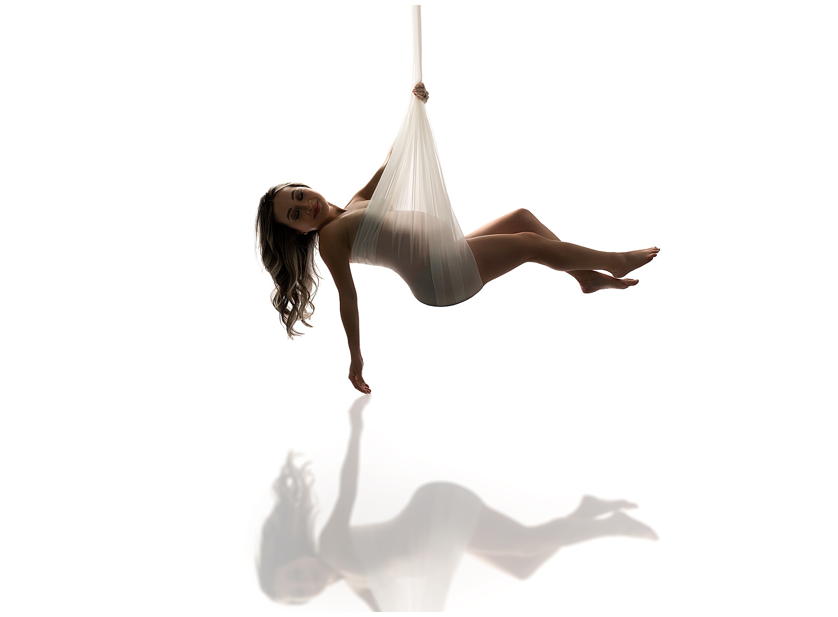 Artistic maternity photo of a pregnant woman in a tulle sling hanging above a neutral, white background. A reflection of the photo is featured beneath. 