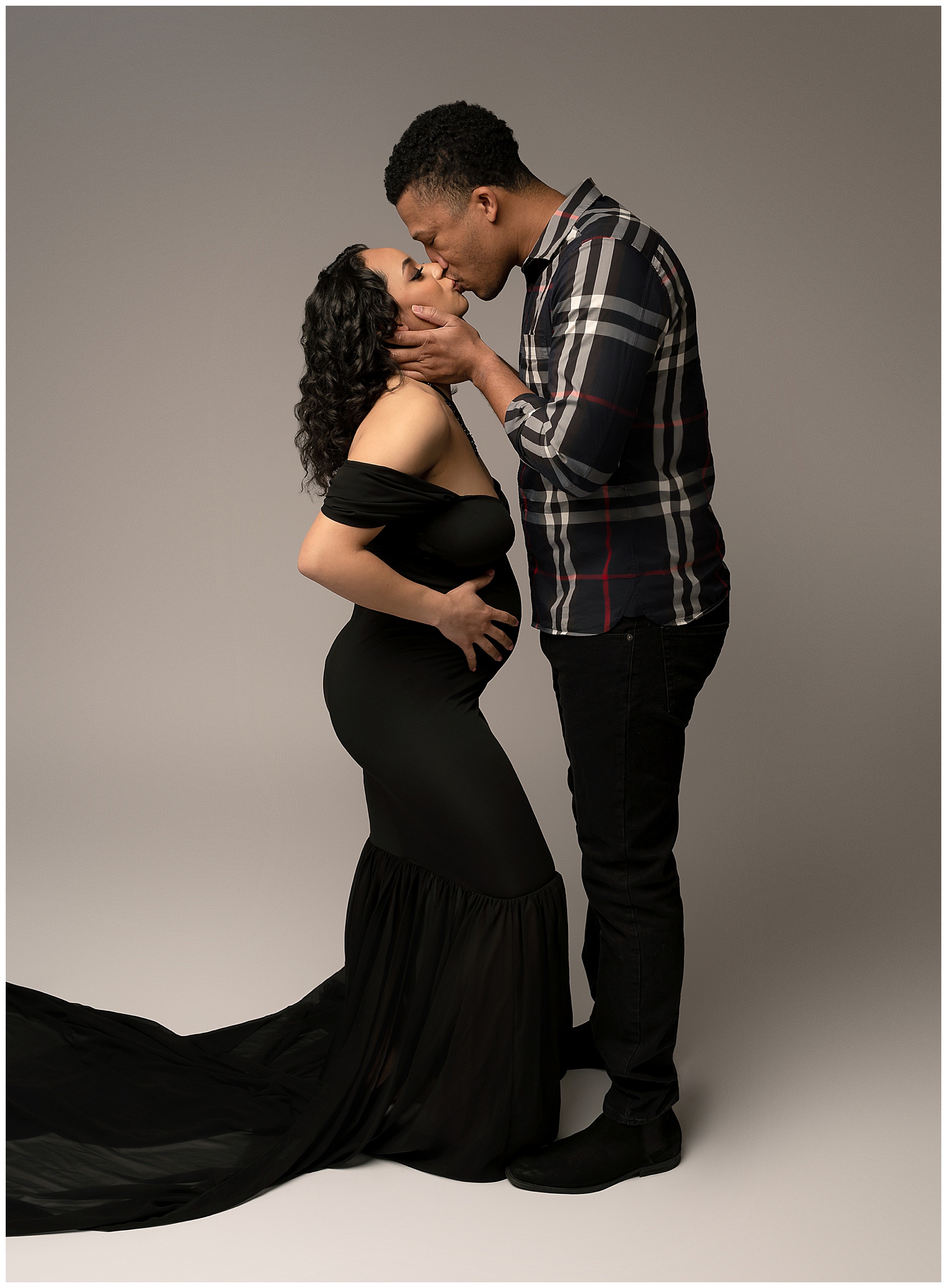 A color maternity photo featuring a pregnant woman and her partner facing each other and kissing. Photo is taken in front of a plain gray background.