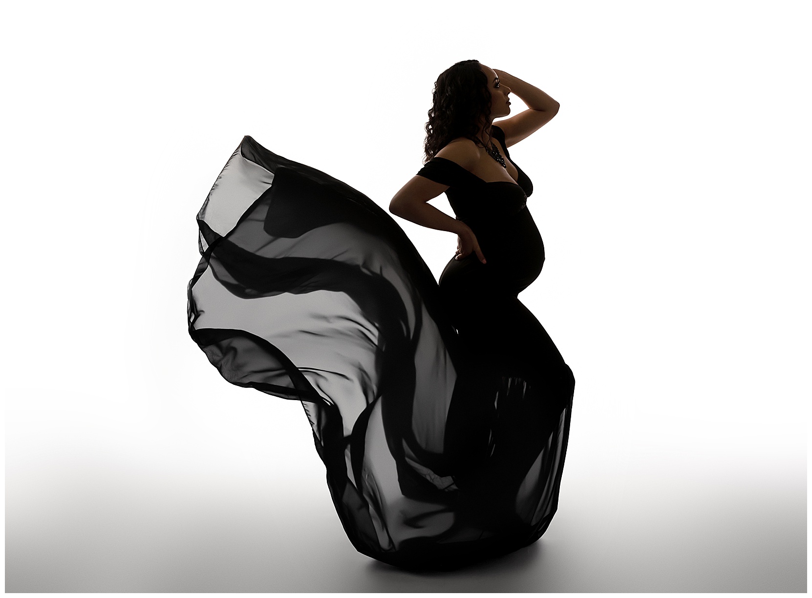 Dramatic baby bump pic silhouette on a white background featuring a woman in shadow wearing a black maternity gown. She is facing toward the right of the picture and the train of her gown is blowing behind her.