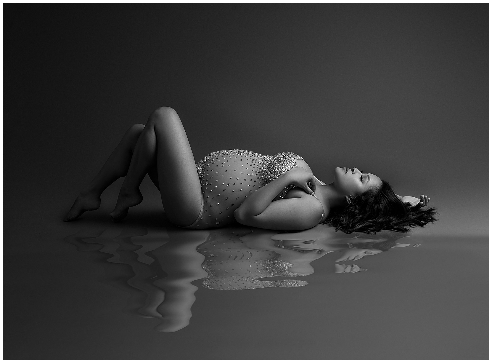 studio maternity photography featuring a woman in a sparkly bodysuit laying on her back. There is a water-like reflection under her.