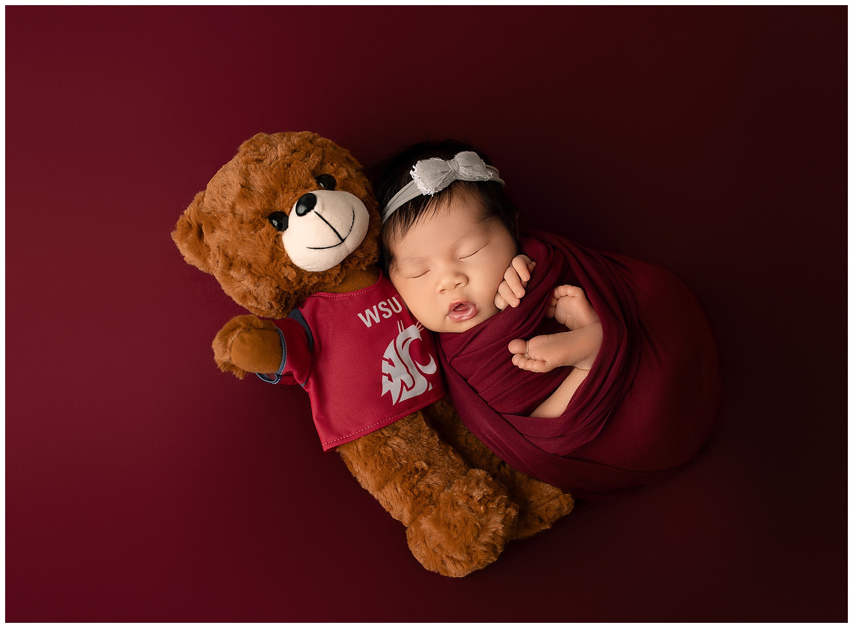 A newborn swaddled in a crimson cloth on a crimson background tucked in next to a teddy bear wearing a Washington State University tee shirt with the Coug logo on the front. 