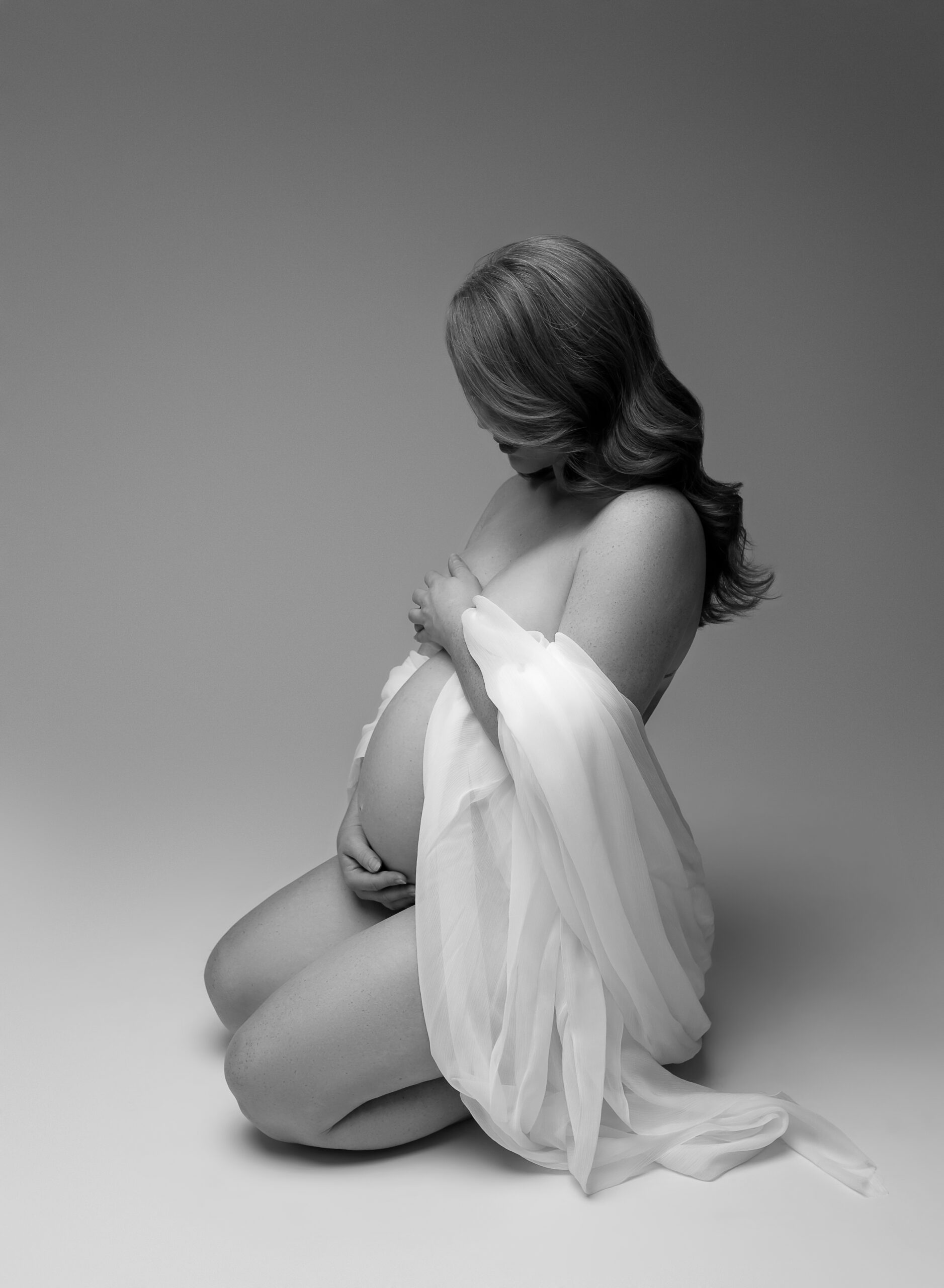 Black and white photo of a pregnant woman on a neutral background. She is kneeling, has white fabric draped around her shoulders, and his holding her baby bump. 