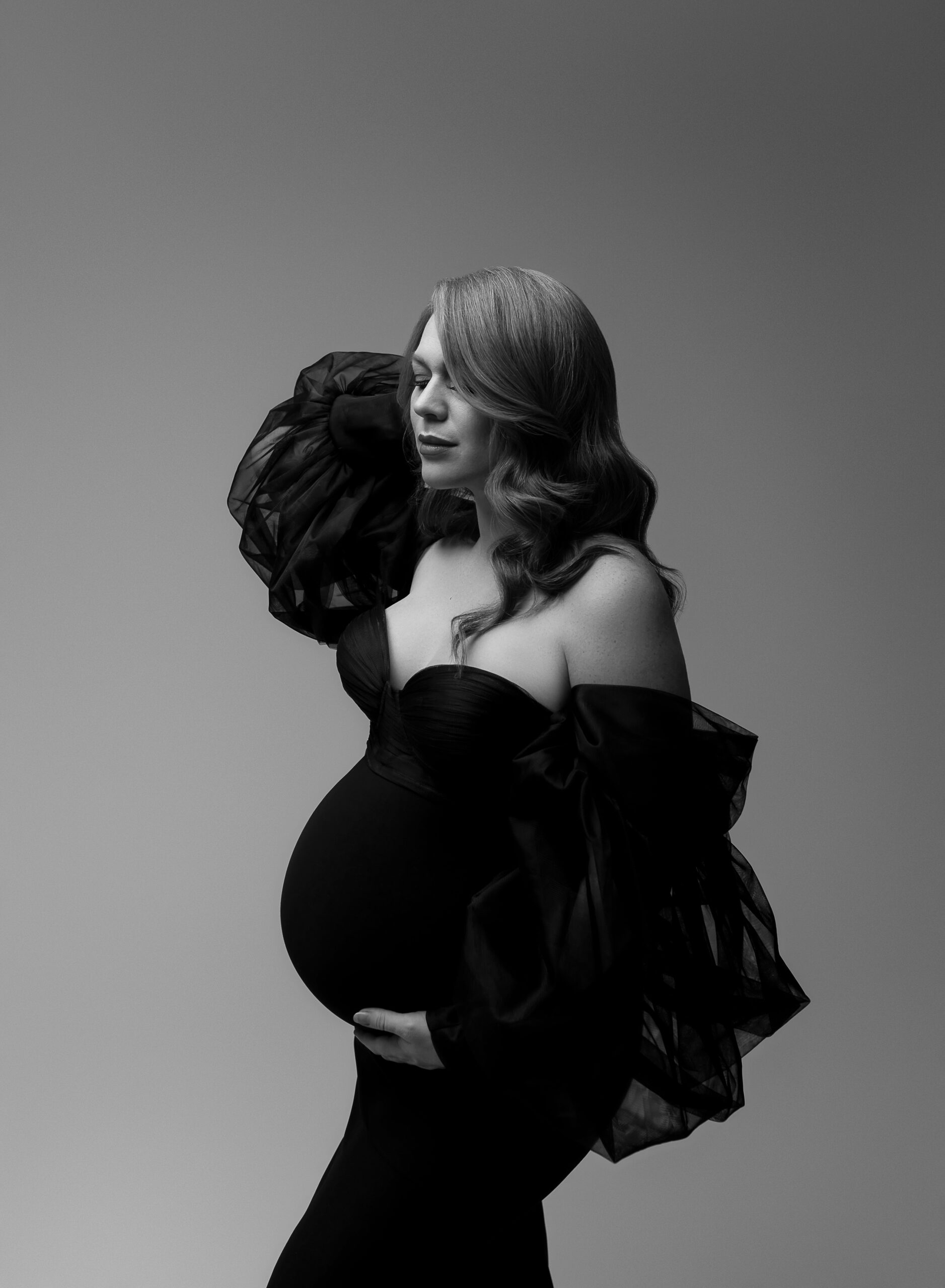 Side profile of a woman in a black maternity gown facing toward the left of the photo. She is holding her baby bump. 