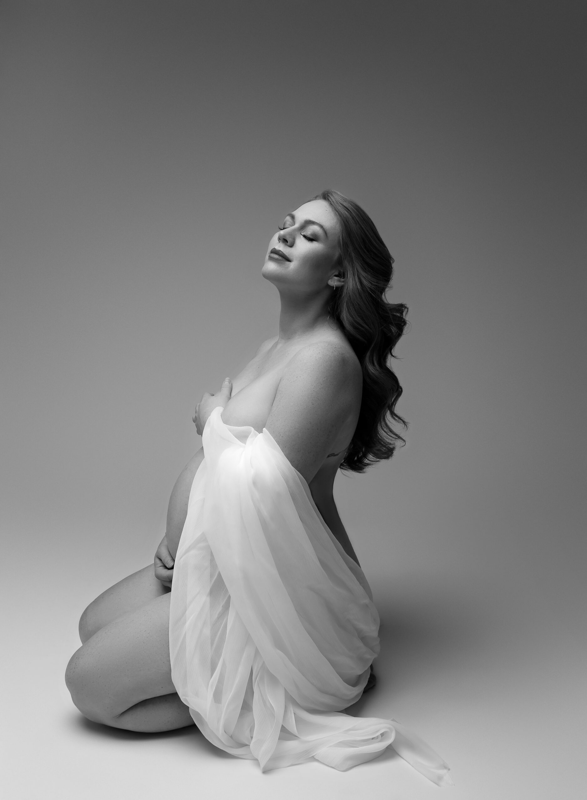 A black and white photo of a pregnant woman kneeling on the ground and looking upward. Her hair falls down her back, and she is wrapped in white fabric.