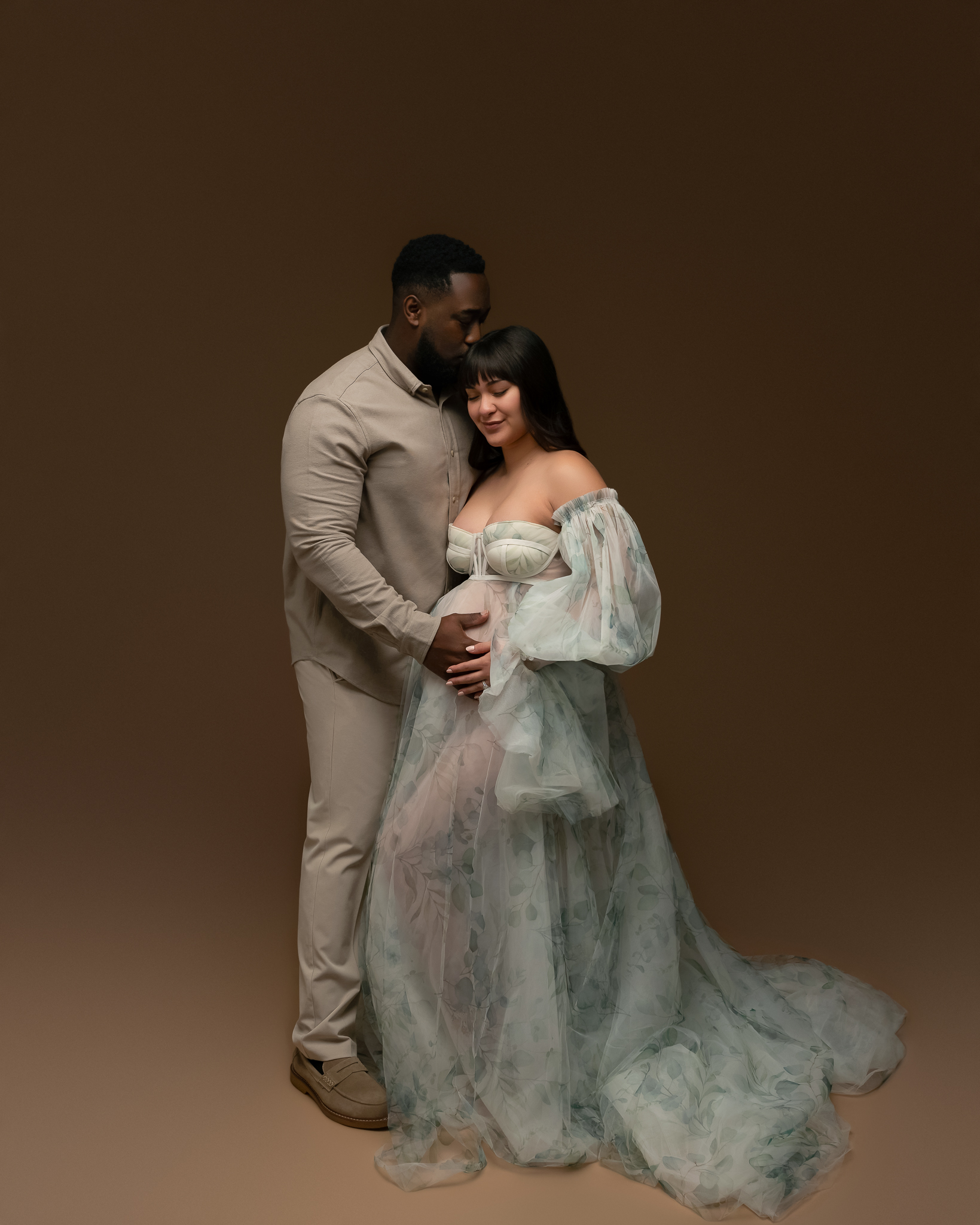 Cedar Park Maternity Studio photo featuring a man and a woman. The man is kissing the woman on the head and they are both holding her baby bump. 