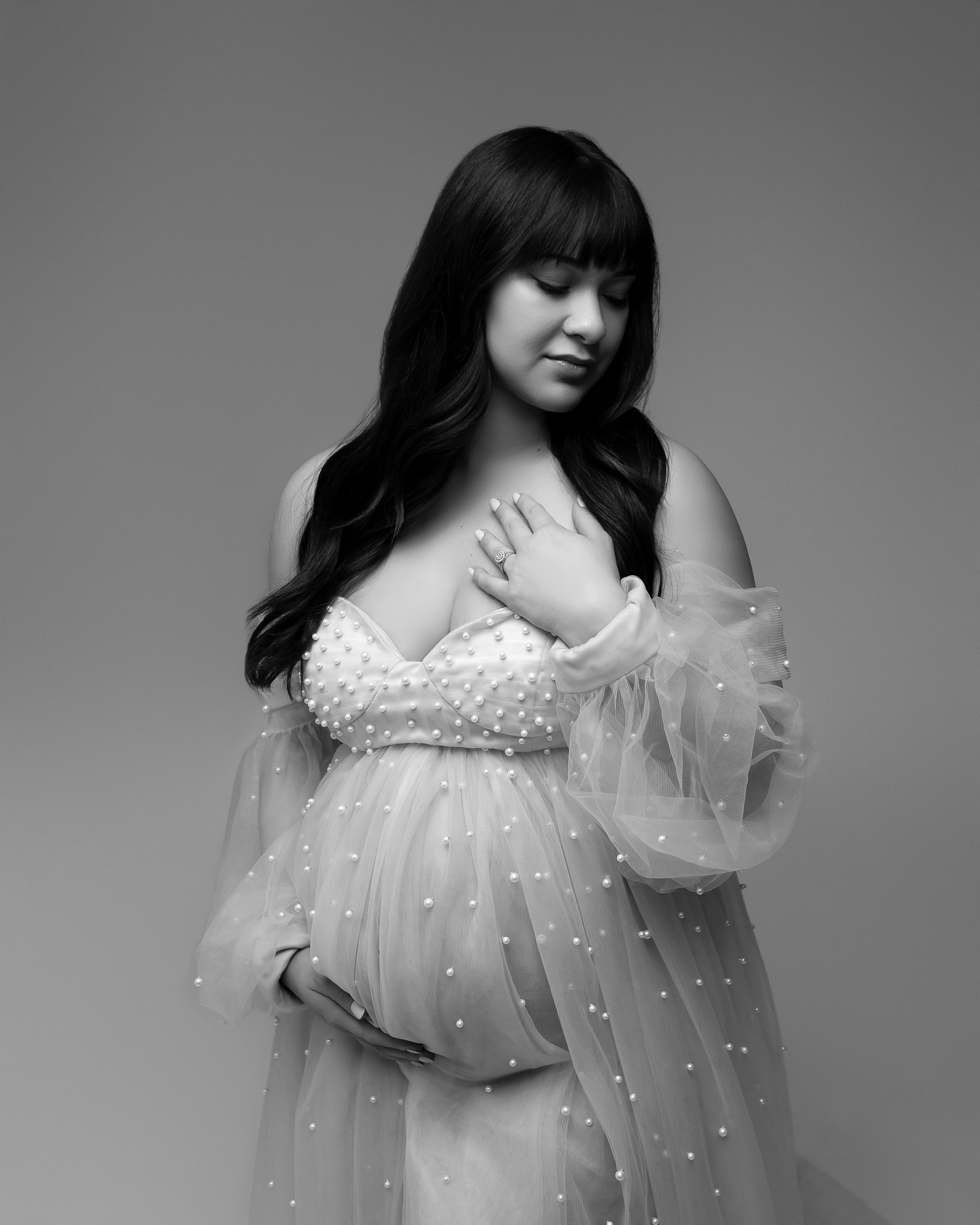 Black and white maternity portrait featuring a woman in a light-colored, sheer maternity gown covered in pearls. 