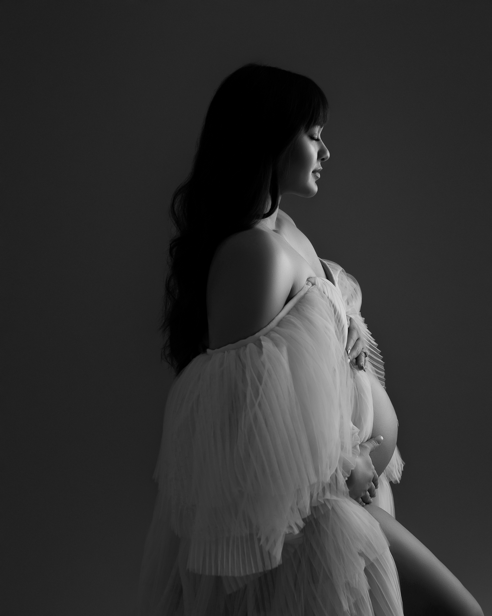 Black and white cedar park maternity studio featuring a woman with long black hair in a frilly, light colored dress. She is facing toward the right of the photo, and light is shining on her face and belly. 