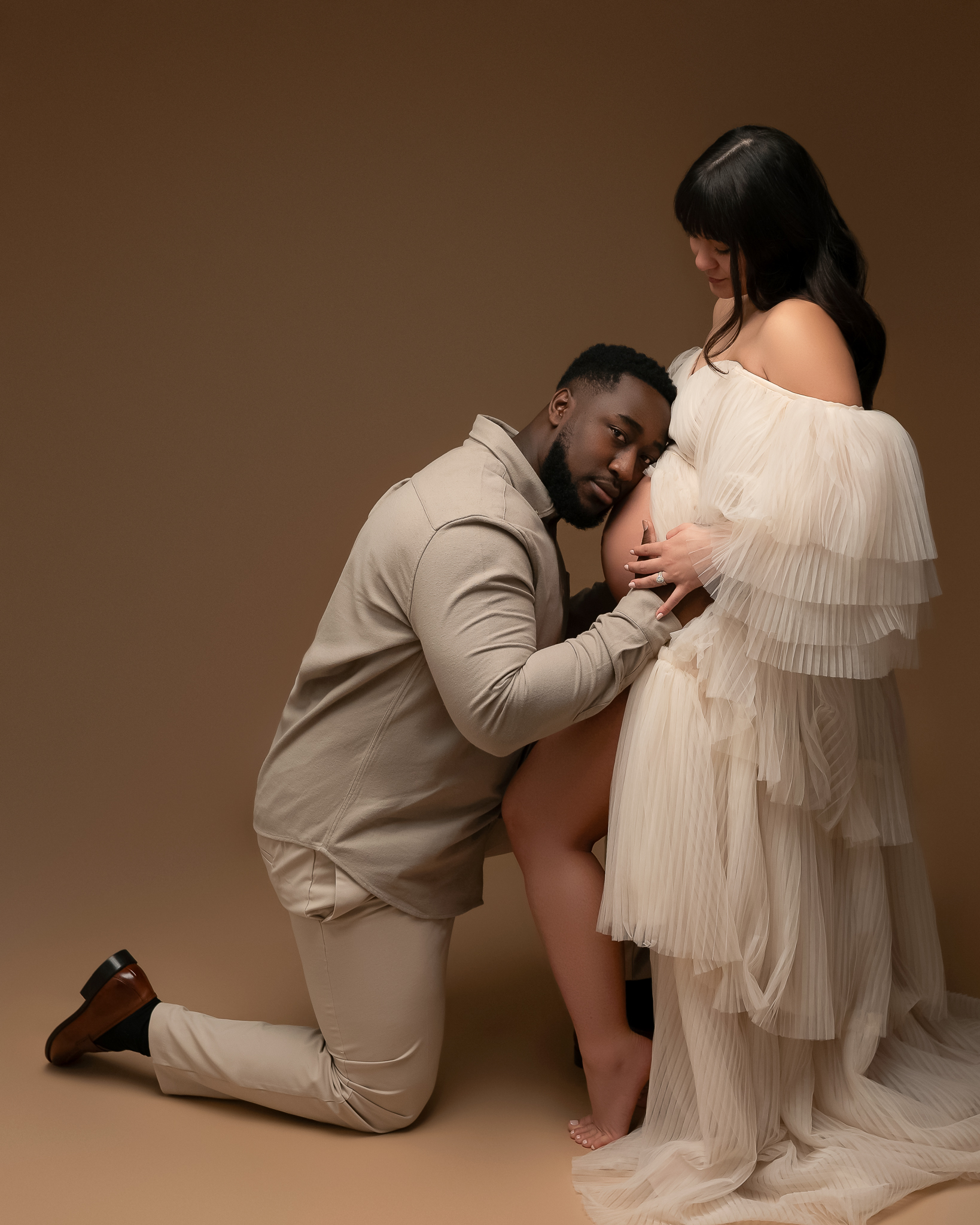 Cedar Park maternity studio photo featuring a man and a woman. The man is kneeling in front of the woman and staring at the camrea with his head on her pregnant belly while she looks down lovingly at him. 