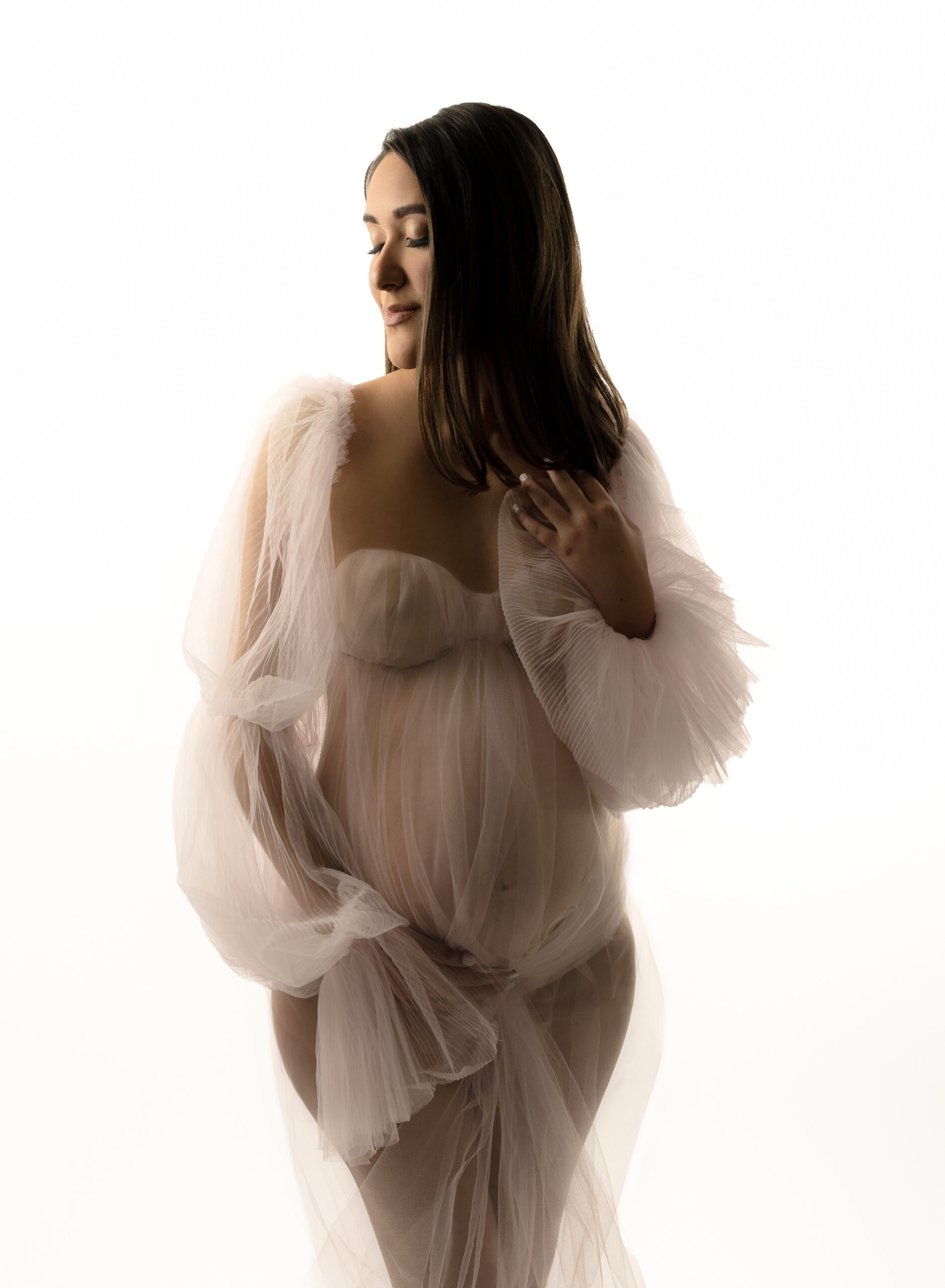 A color photo featuring a pregnant woman wearing a sheer tulle maternity gown. She has one hand on her collarbone, one holding her belly, and she is looking down with her eyes closed.