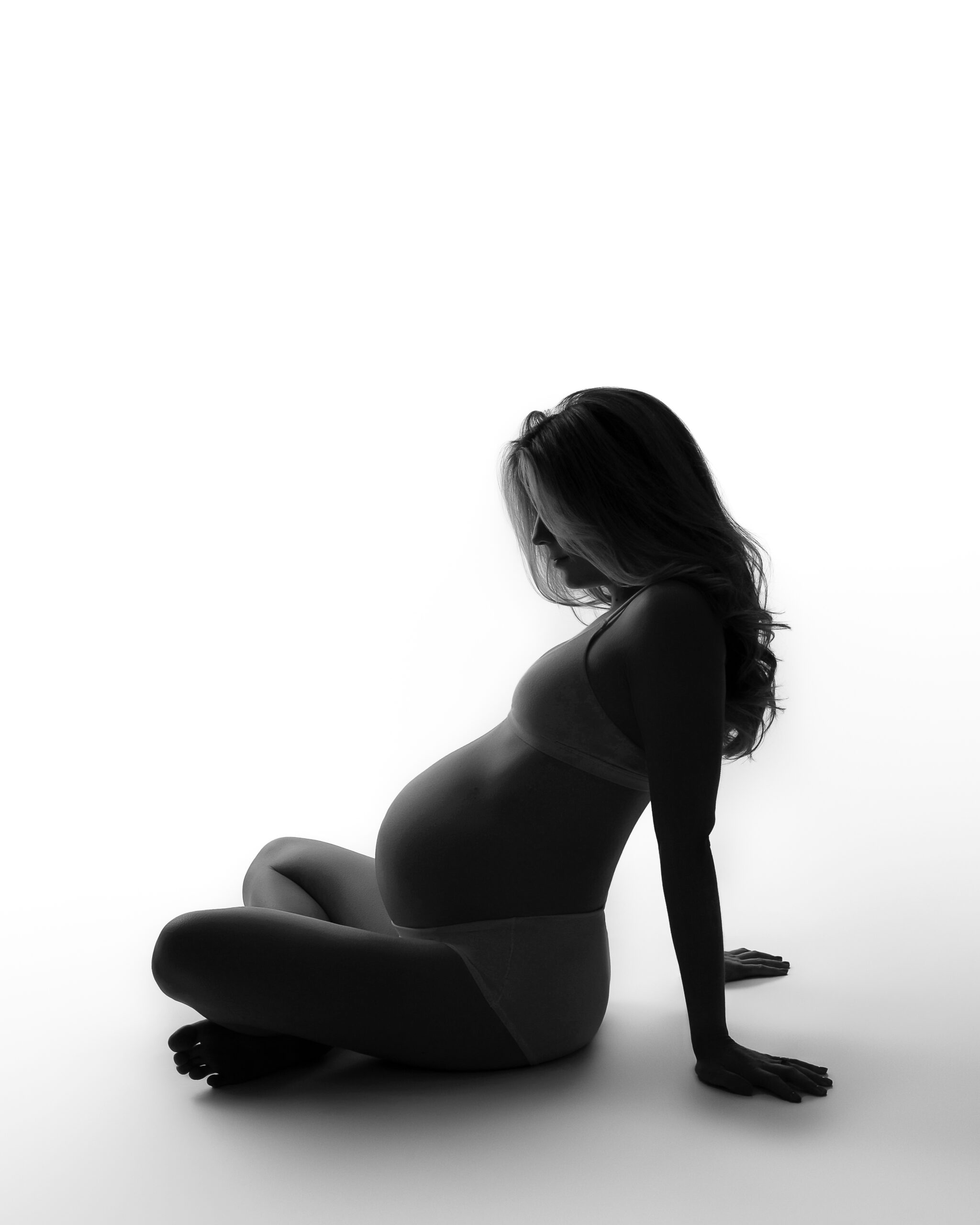 A black and white photo of a pregnant woman in shadow on a white background. She is sitting cross-legged and leaning back, emphasizing her belly. 