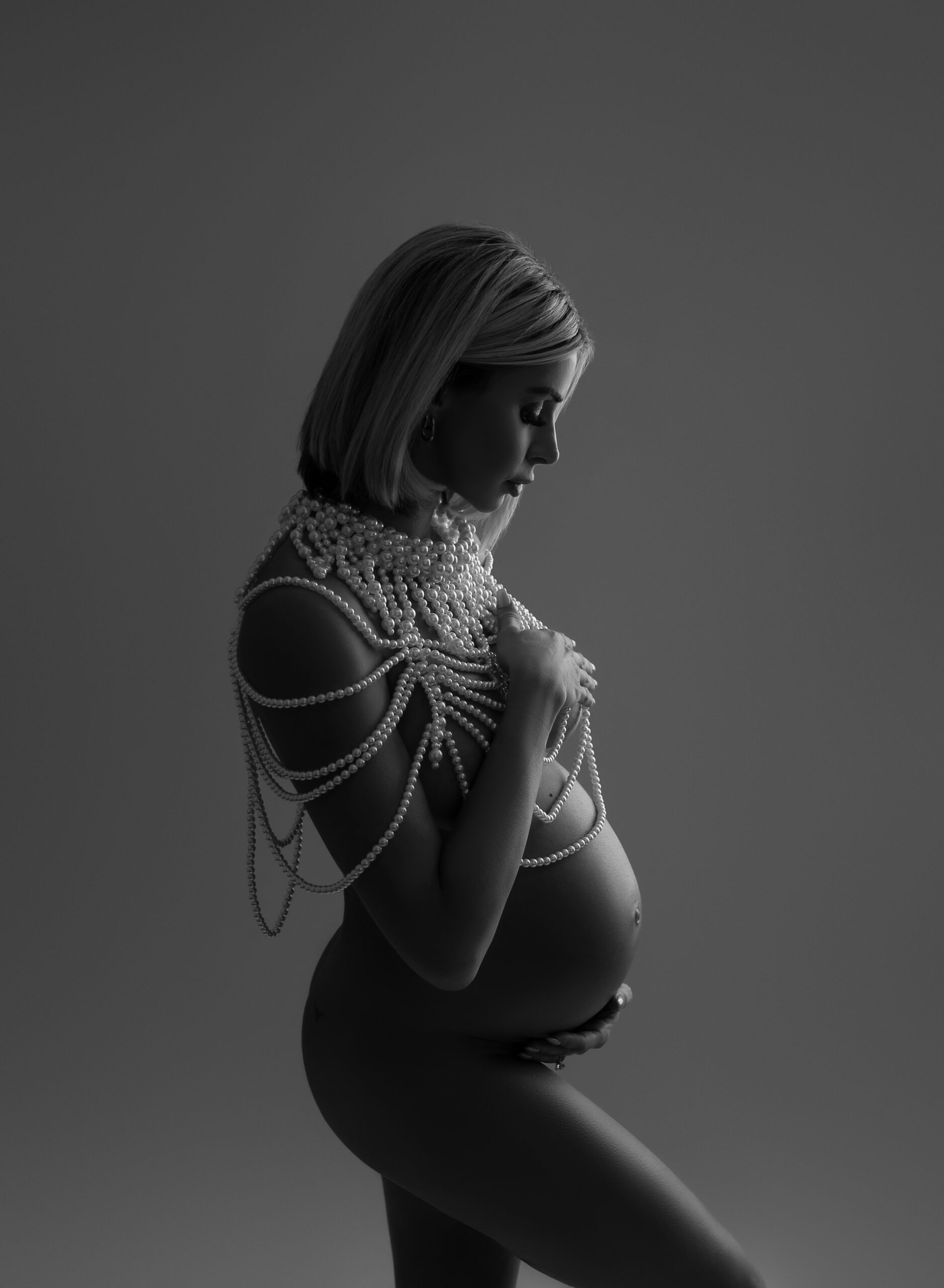 A black and white boudoir maternity photo featuring a woman with short blonde hair covering her chest and holding her baby bump. She is naked and covered in pearls on the upper part of her body. 