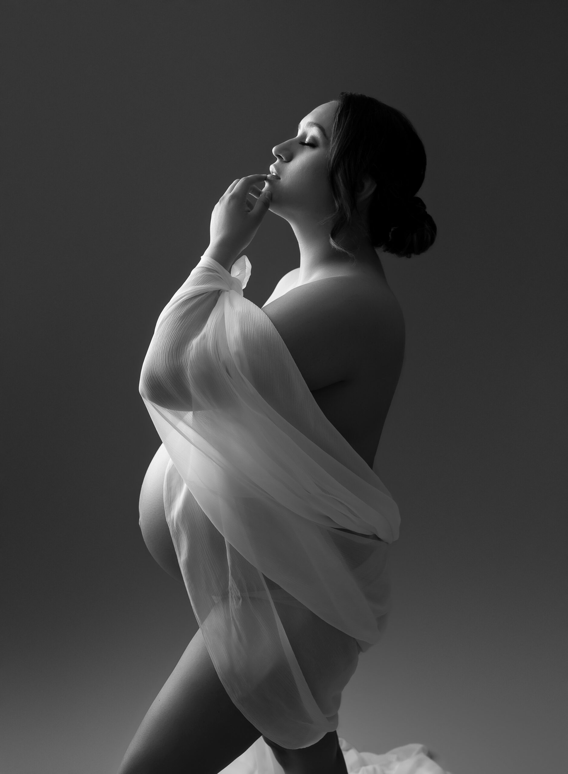 A nude pregnant woman stands in profile faced to the left. She is draped in lightly-colored tulle and holds her hand to her lips. 