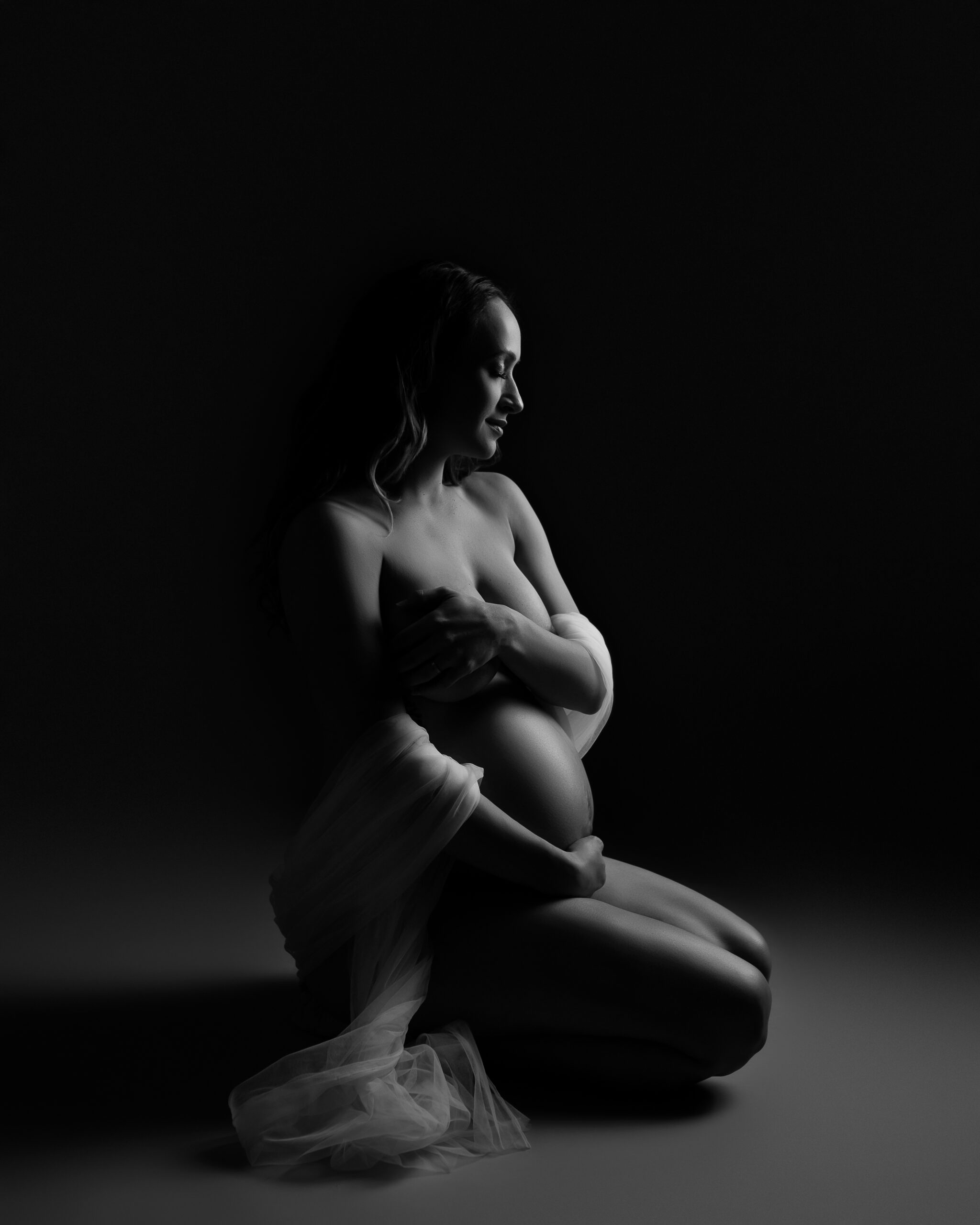 A black and white boudoir photo featuring a pregnant woman in soft lighting kneeling on the floor. She is lightly draped in tulle across her arms and back, and is tastefully covering her breasts and holding her belly.