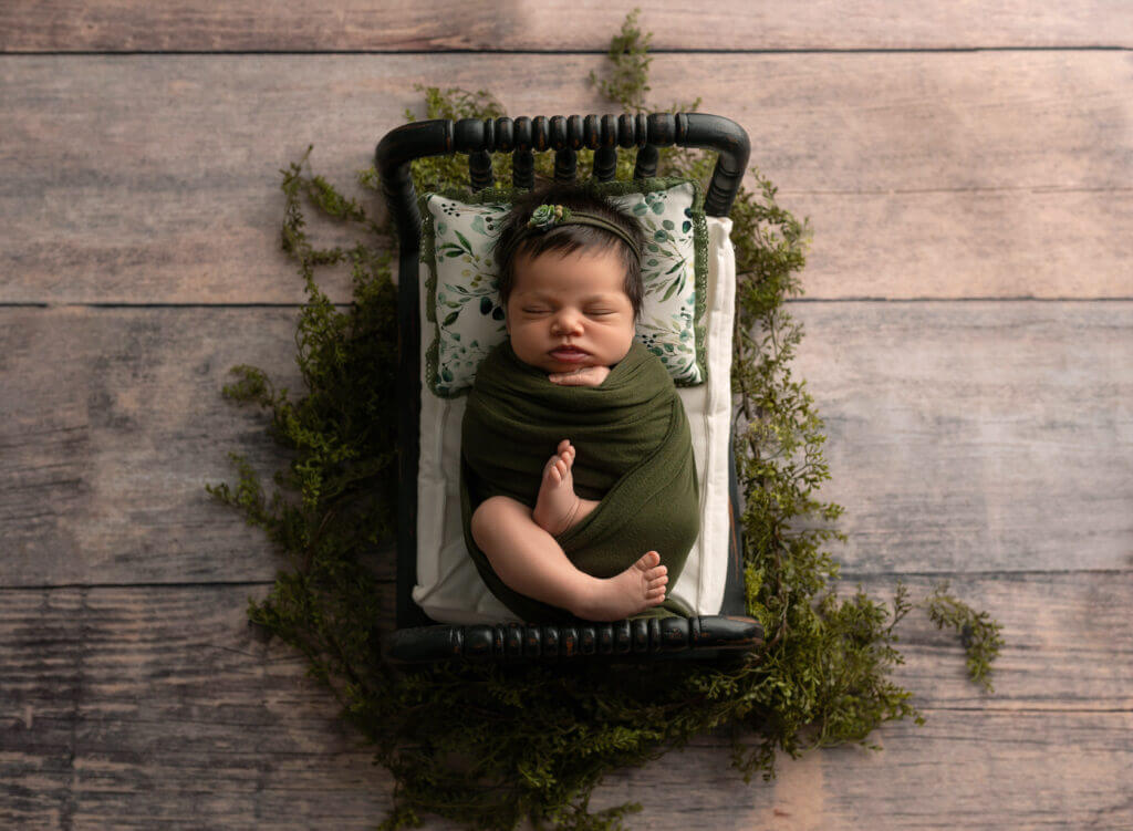 Newborn wrapped in olive green knit blanket lying in vintage black metal baby bed with leaf-patterned pillow surrounded by natural moss and greenery on weathered wooden floor