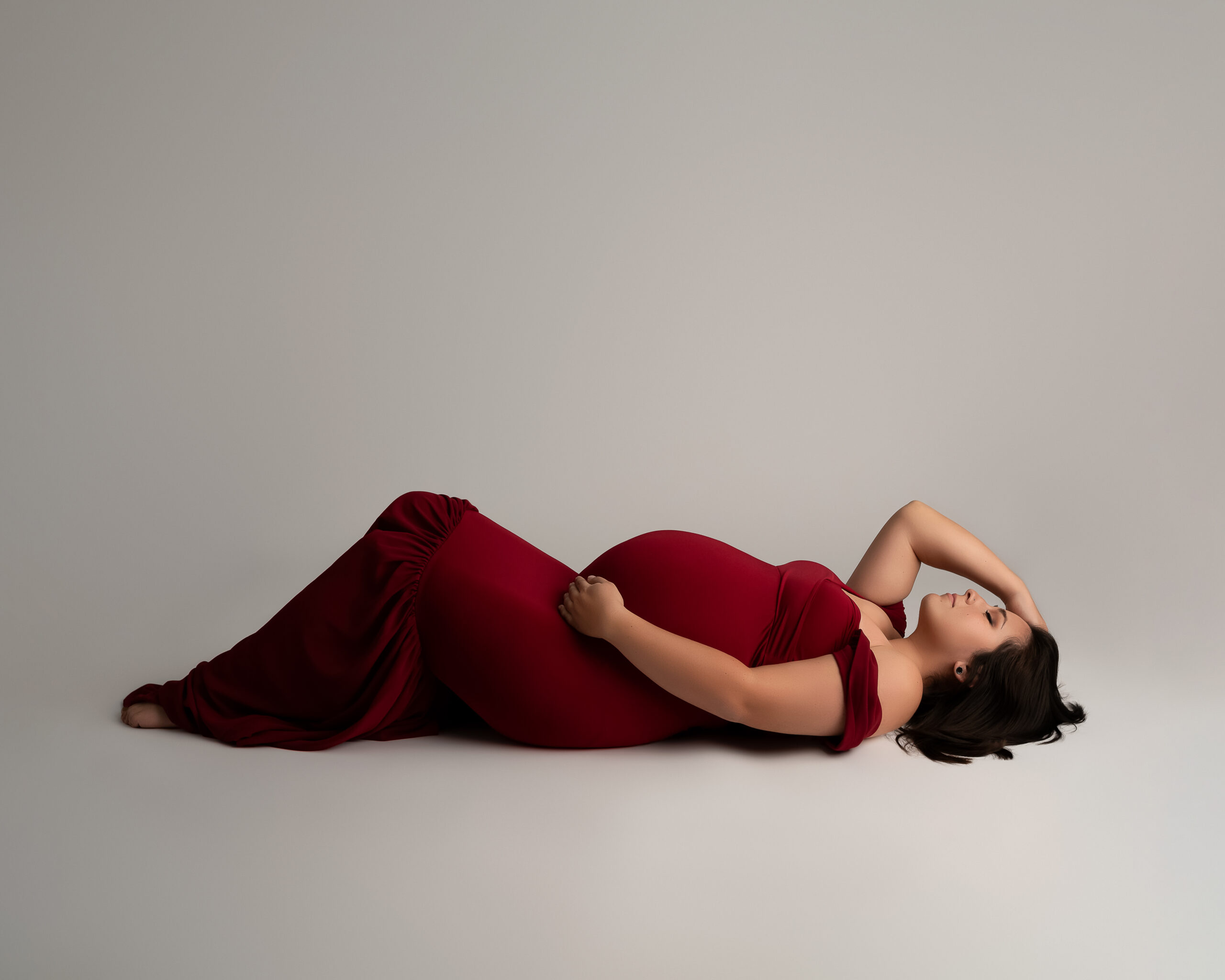 Woman in a red maternity gown is laying on her back on the floor. She is cradling her baby bump and has her hand on her head. 