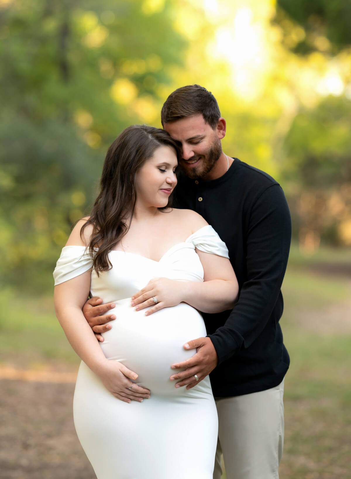 A man stands behind a woman on a wooded path. They are both holding the woman's pregnant belly. 