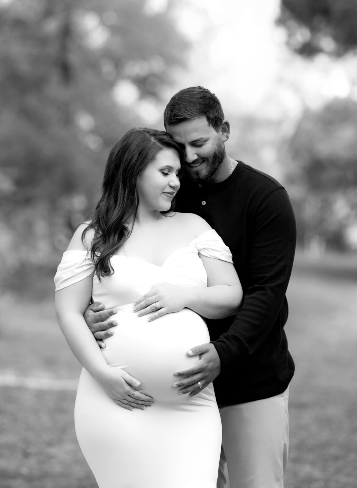 A black and white photo of a man and woman on a wooded path. The man is standing behind the woman and they are both holding her pregnant belly. 