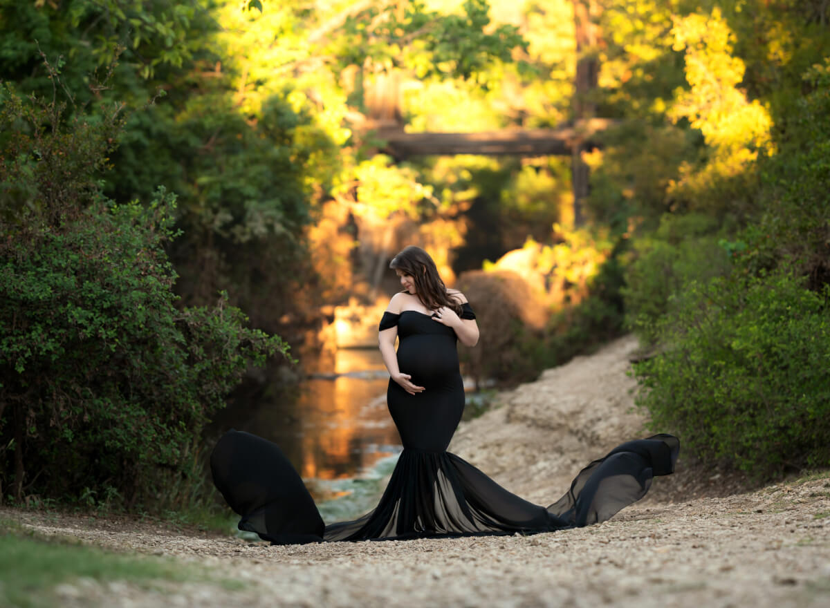 Sunrise maternity photos at Brushy Creek, pregnant woman in a flowy black gown holding her bump beside the creek