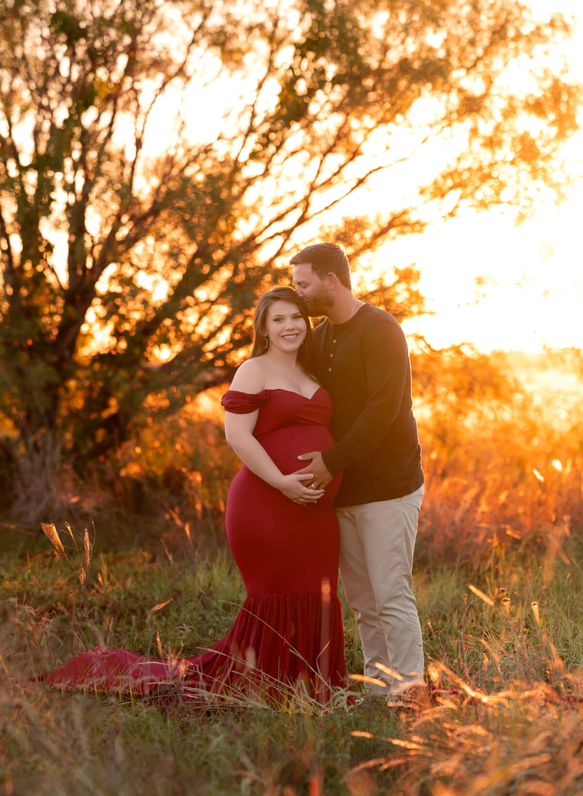 A pregnant woman in a red maternity gown and a man in a black shirt and khakis stand in a wooded area in front of the rising sun. He is holding and kissing her on the head, and they both have one hand on her stomach.