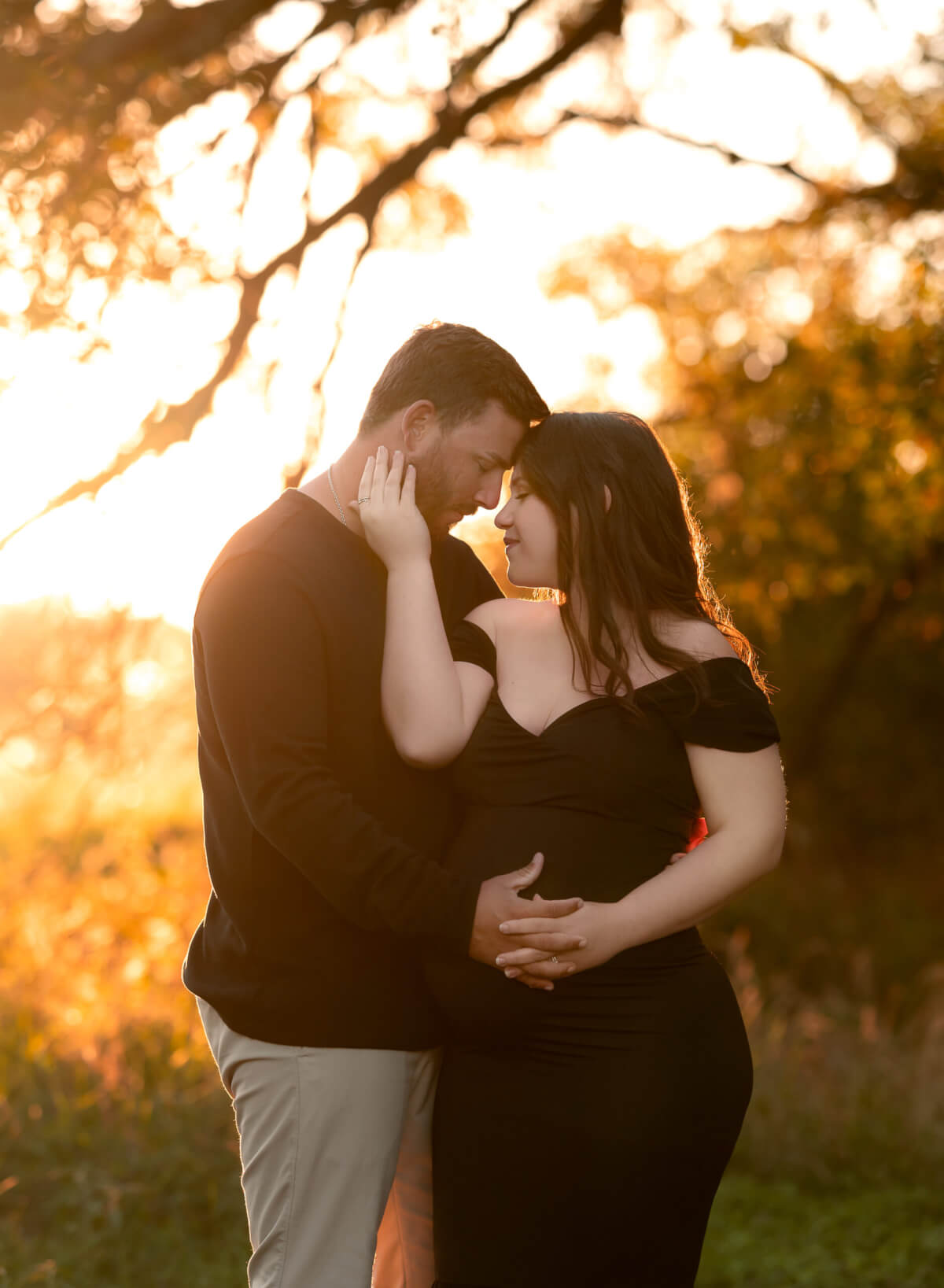 A pregnant woman in a black gown and a man in a black shirt are standing side by side with their foreheads pressed together. The sun is rising behind them.
