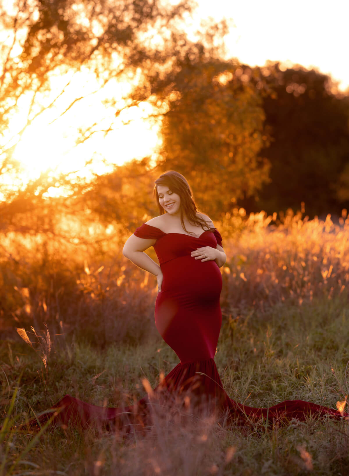 A pregnant woman stands in a field in Brushy Creek park as the sun rises behind her. She is wearing a red maternity gown and looking and smiling with one hand on her hip and the other on her stomach.