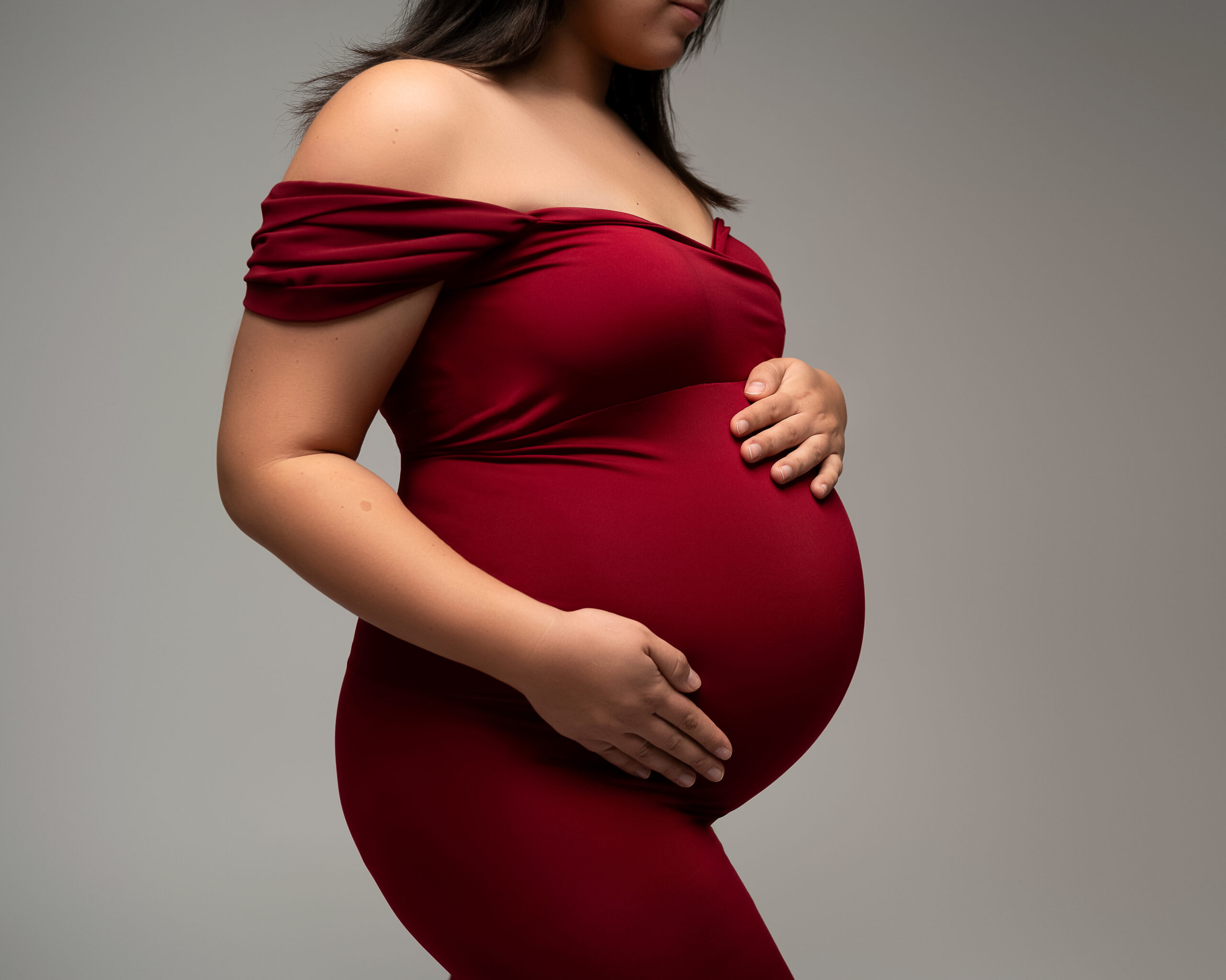 Maternity photo featuring a woman in a red maternity gown looking down at her baby bump. 