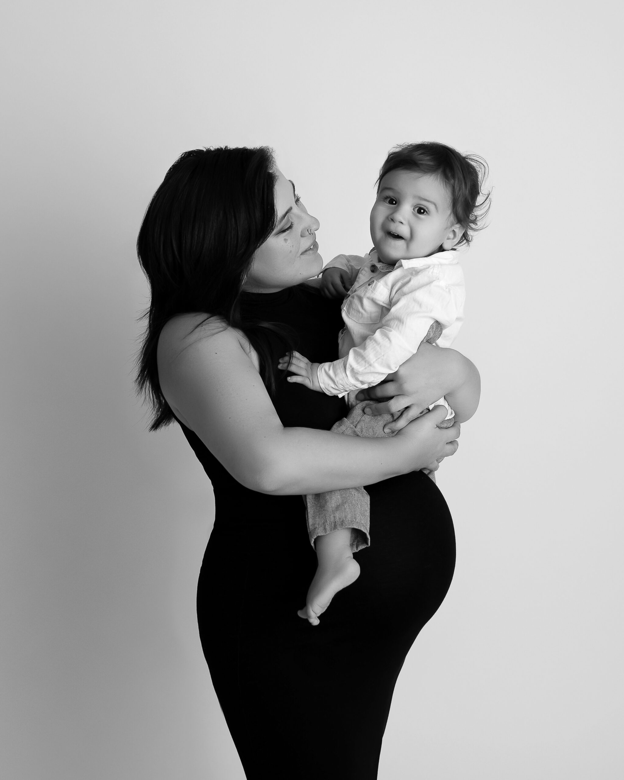 Black and white maternity photo featuring a pregnant mom in a black maternity gown holding her toddler son. 