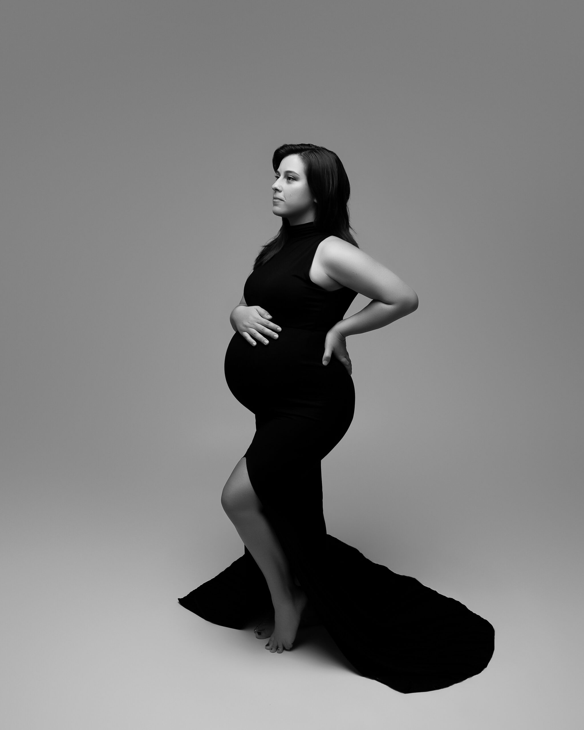 Full body maternity portrait featuring a woman in a black maternity gown facing the left of the photo. She has her hand on her baby bump and is looking wistfully off camera.