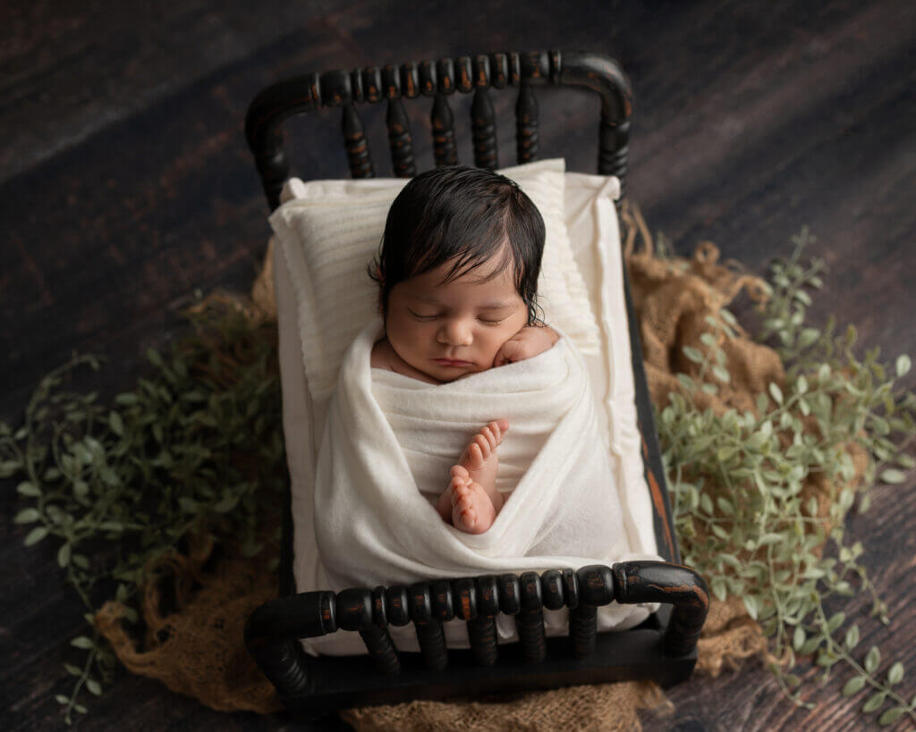 Newborn wrapped in cream knit blanket sleeping in vintage black metal bed frame surrounded by natural moss and eucalyptus branches on dark rustic wooden floor
