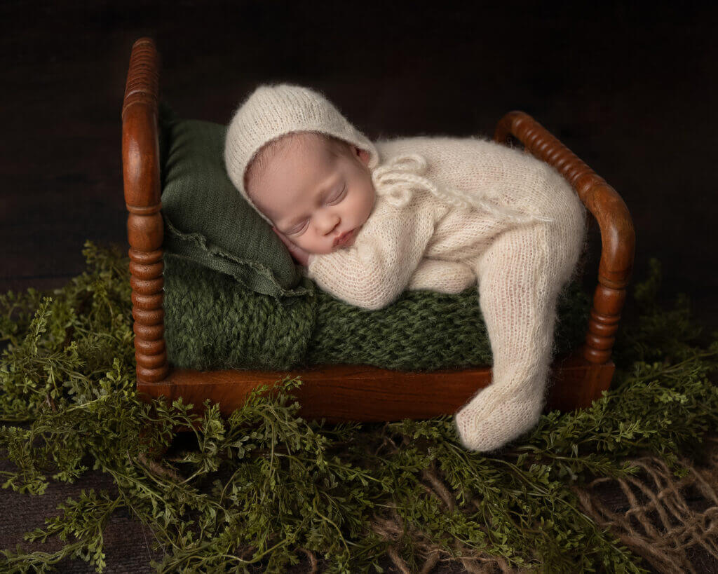 Newborn baby in cream cable-knit outfit and matching bonnet lying on moss-covered wooden bench surrounded by natural woodland foliage and dried branches