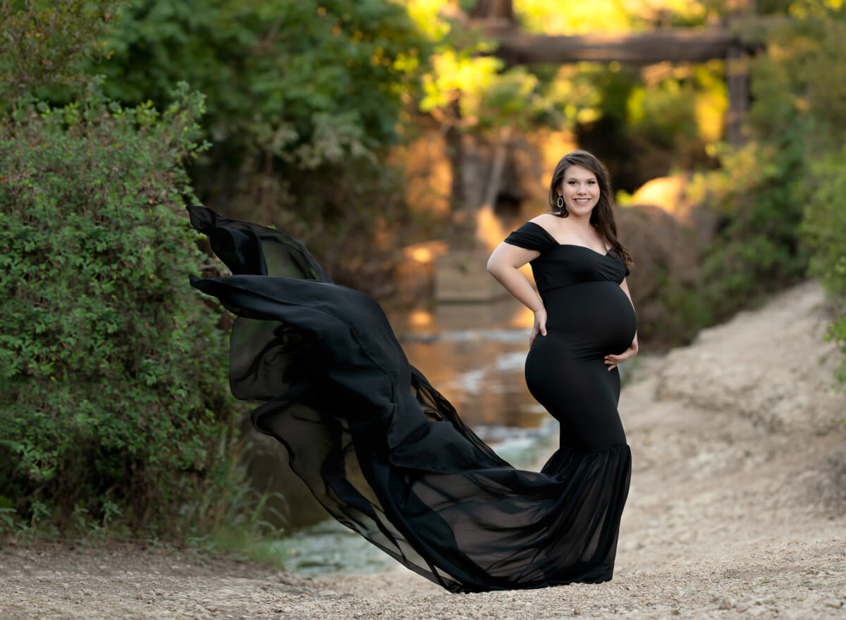 A woman in a black maternity gown stands in front of Brushy Creek in the Austin, TX area. She is standing profiled to the side with one hand on her tummy and one on the small of her back.