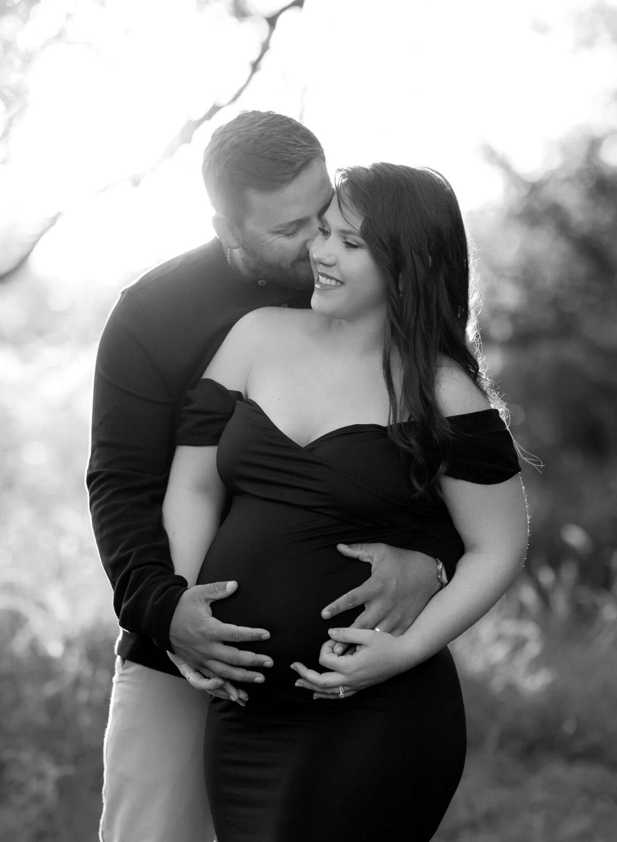 A black and white maternity photo featuring a man holding a pregnant woman from behind. She is smiling, and sun is beaming behind them as they both hold her baby bump. 