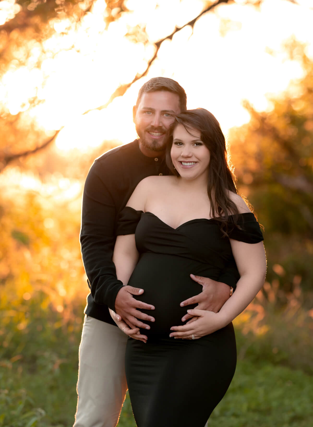 A man and woman stand together in front of a sunrise. They are looking directly at the camera and holding her pregnant belly. 