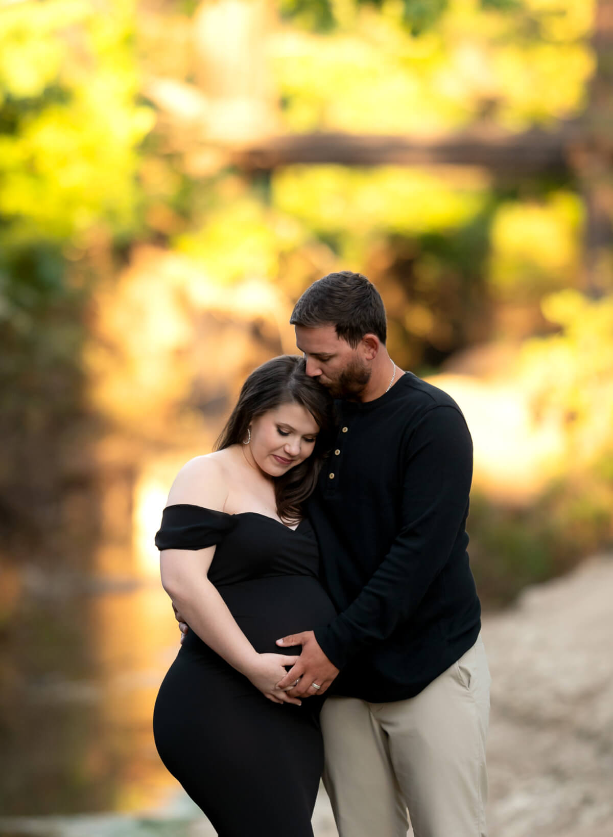A man and a woman stand in front of a wooded area in Brushy Creek park. She is in a black maternity gown, he is in a black Henley shirt, and they are both holding her baby bump.